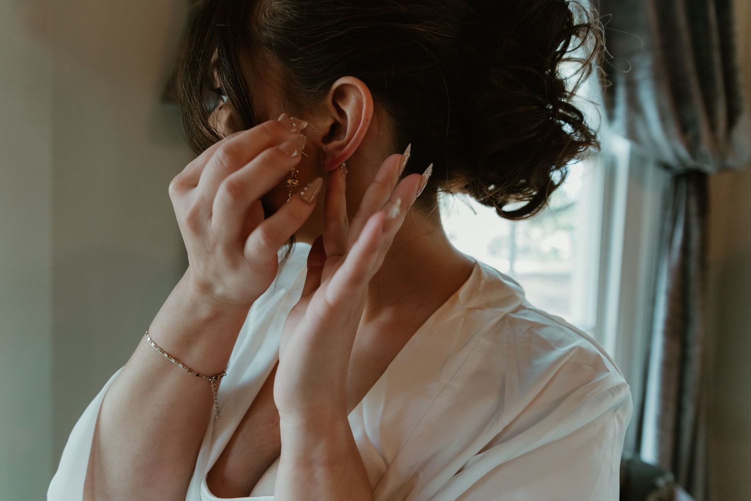 Bride in white robe putting on earrings while getting ready on her wedding day.