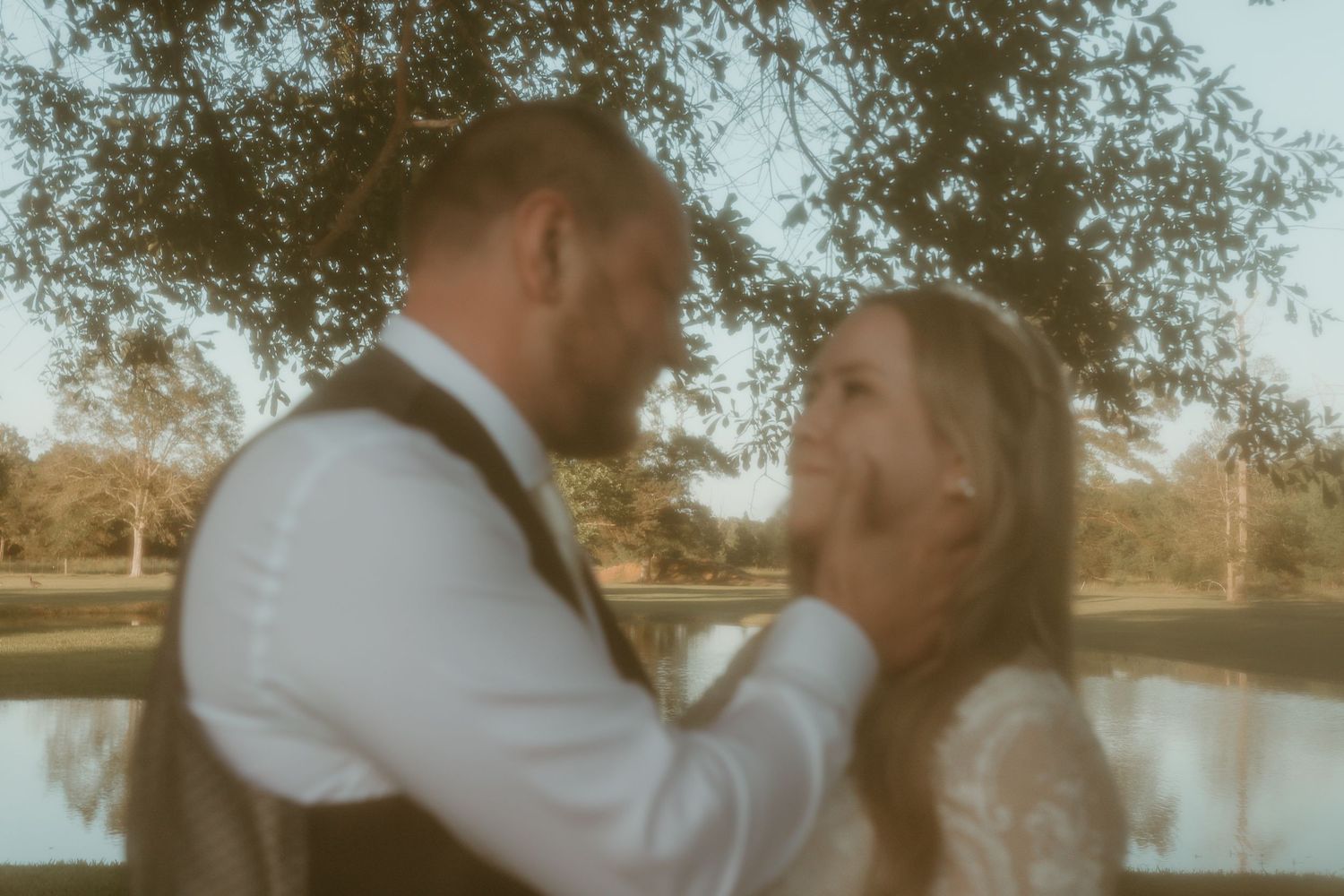 Bride and groom share a close romantic moment beneath a tree with a peaceful pond in the background.