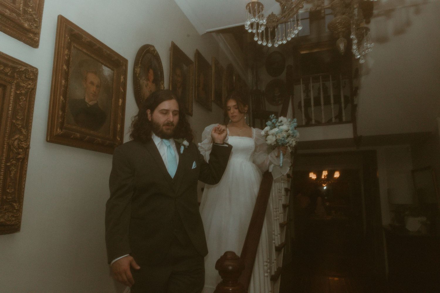 Groom leads bride down elegant staircase lined with portrait paintings in a historic mansion.