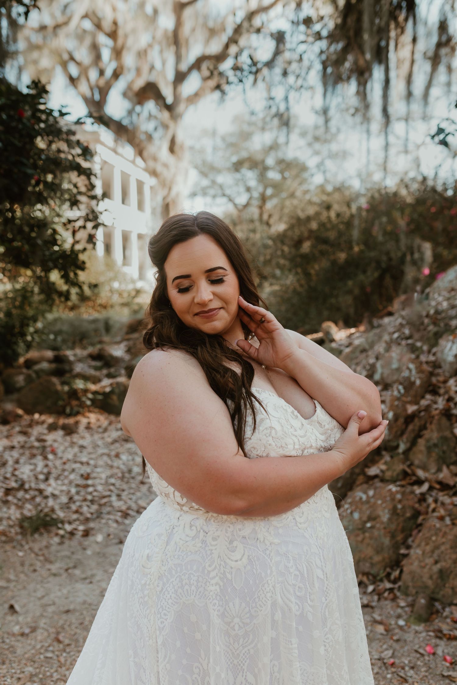 Bride in lace A-line wedding gown touching earring, posing in dappled light near stone wall.
