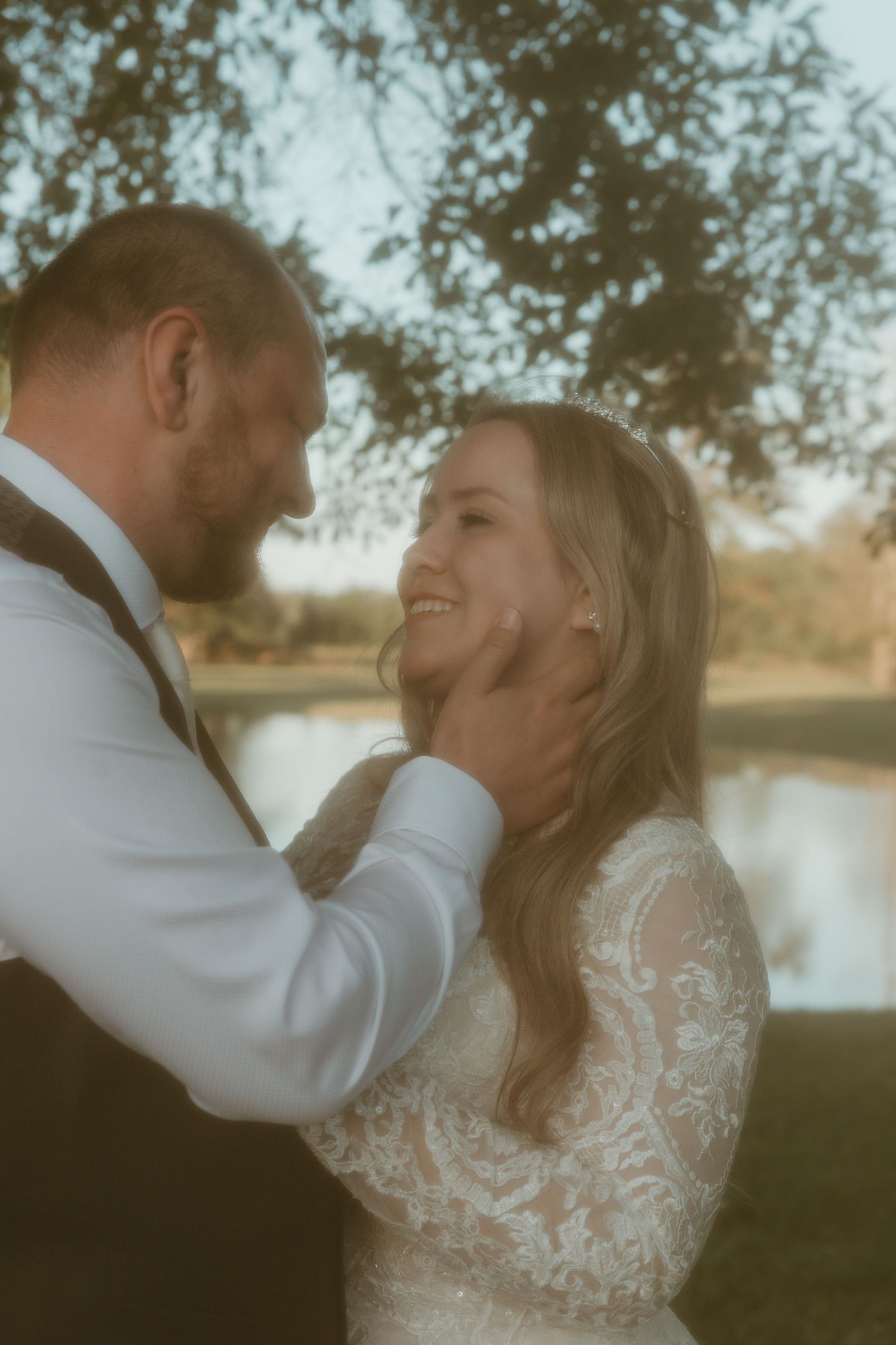 Groom tenderly holds brides face as she smiles wearing a lace gown and tiara by a pond.