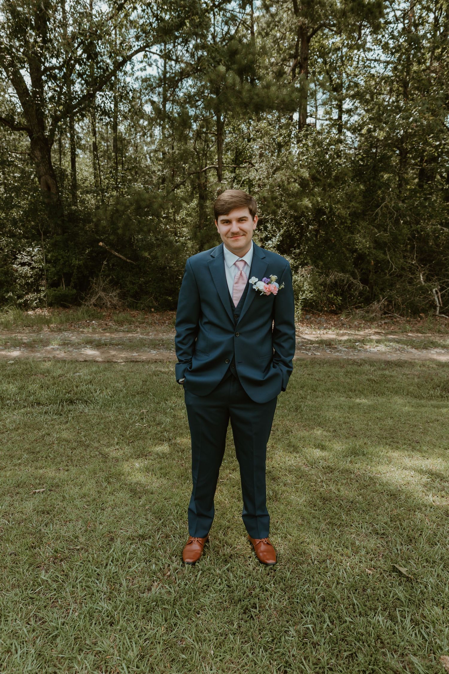 Groom in navy three-piece suit with floral boutonniere stands on grass with hands in pockets.