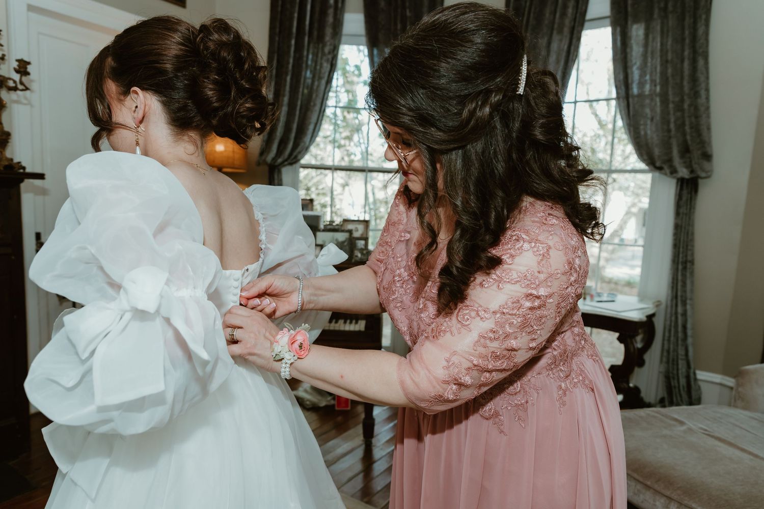 Mother helping bride button up her white puff sleeve wedding dress in a beautifully decorated bridal suite.