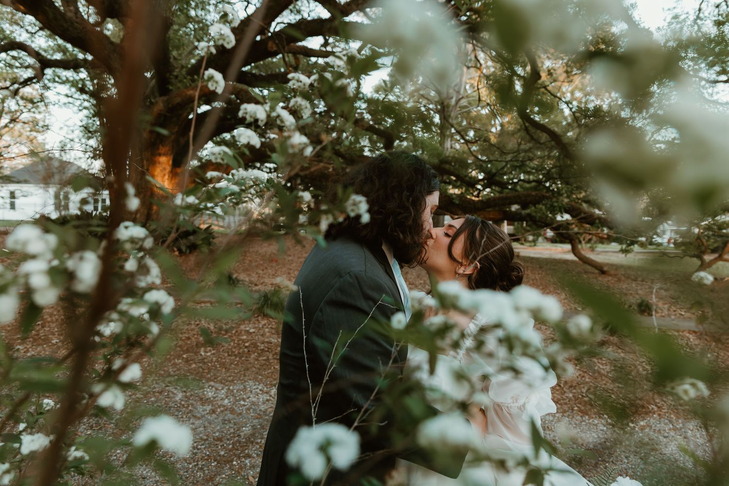 Couple shares romantic kiss among blooming white flowers in a garden during their wedding day.