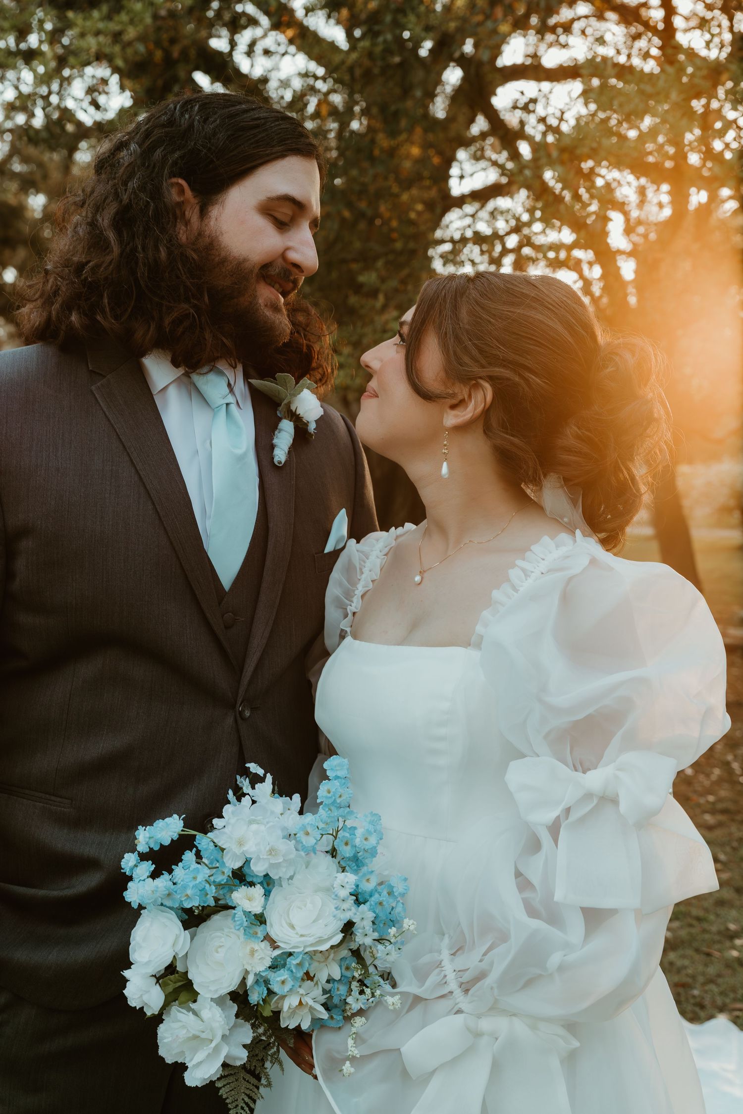 Bride and groom share a golden sunset moment, bride holding blue and white bouquet.