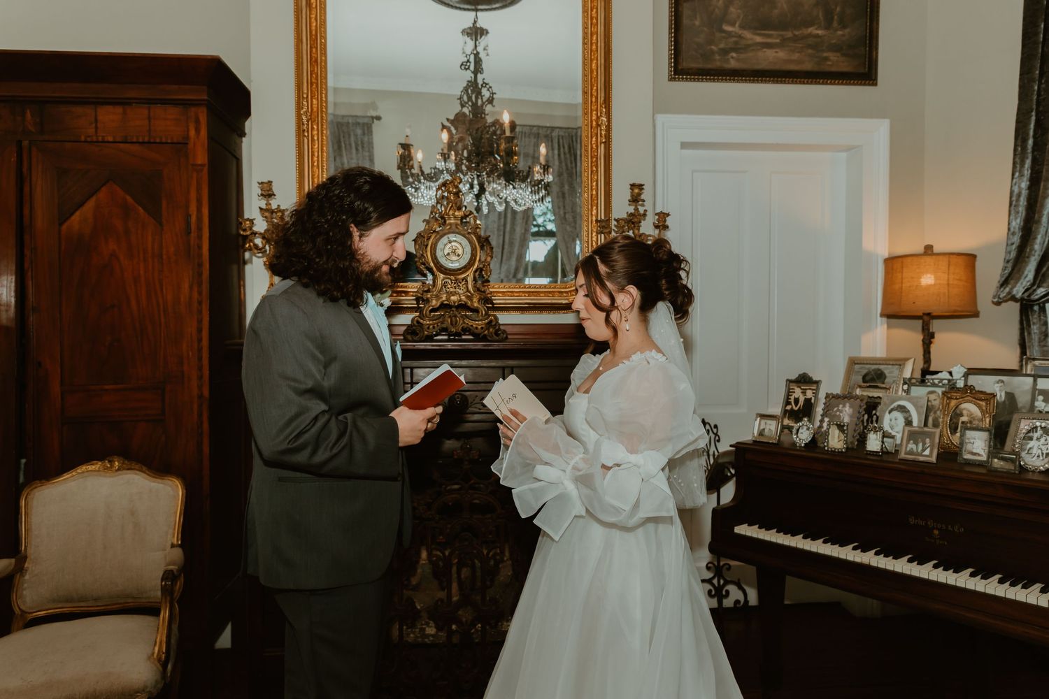 A bride and groom exchange handwritten vows indoors beside a grand piano and ornate fireplace mantel.