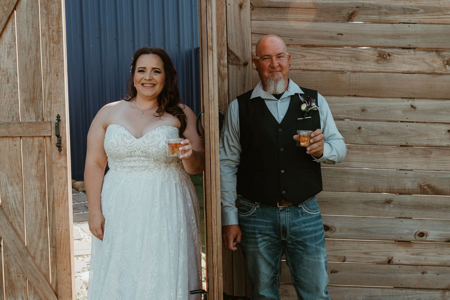 Bride and groom smiling while holding whiskey glasses in front of rustic wooden barn doors.