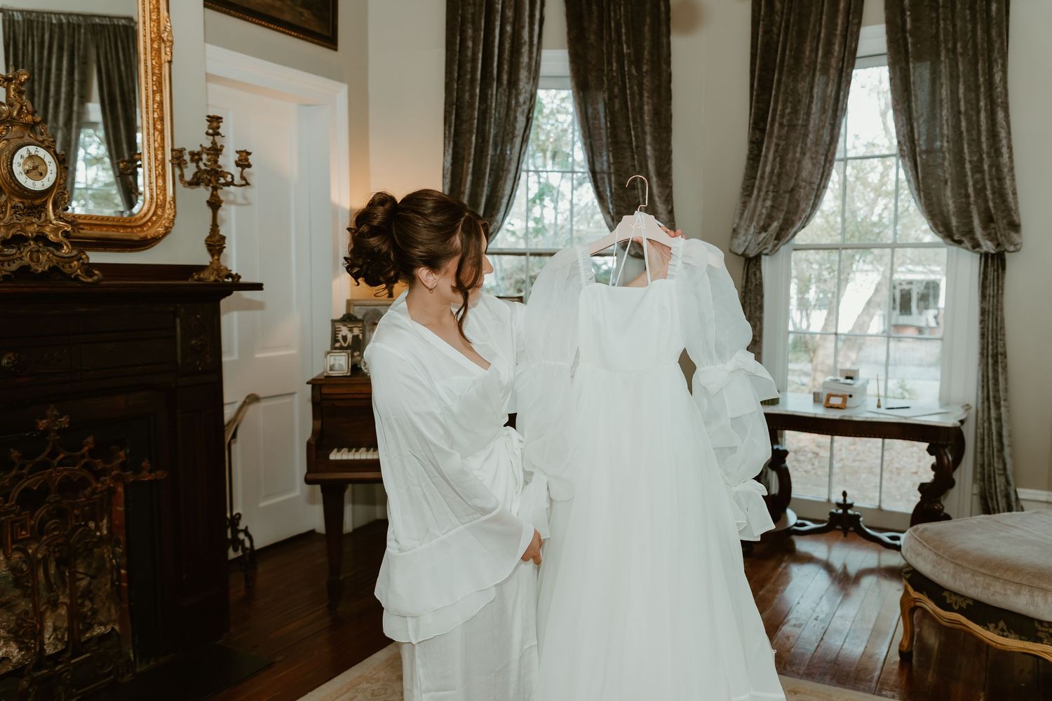 Bride in white robe admiring her wedding gown hanging in an elegant room with fireplace and chandelier.