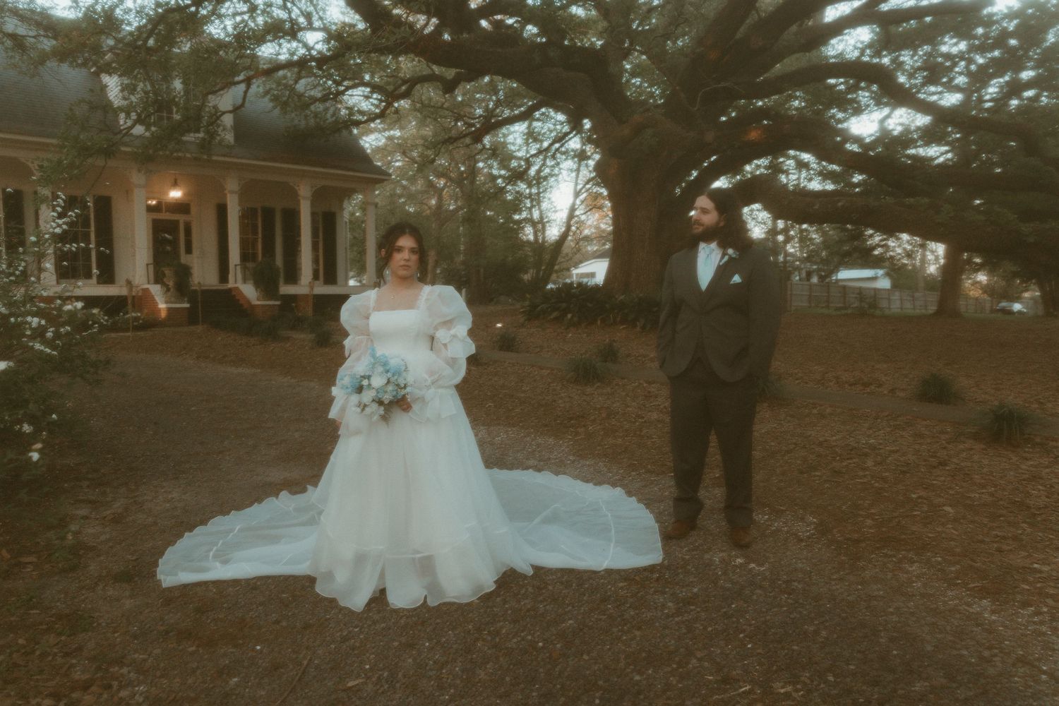 Bride in white ballgown and groom in gray suit stand apart outdoors near large tree at elegant Southern estate wedding.