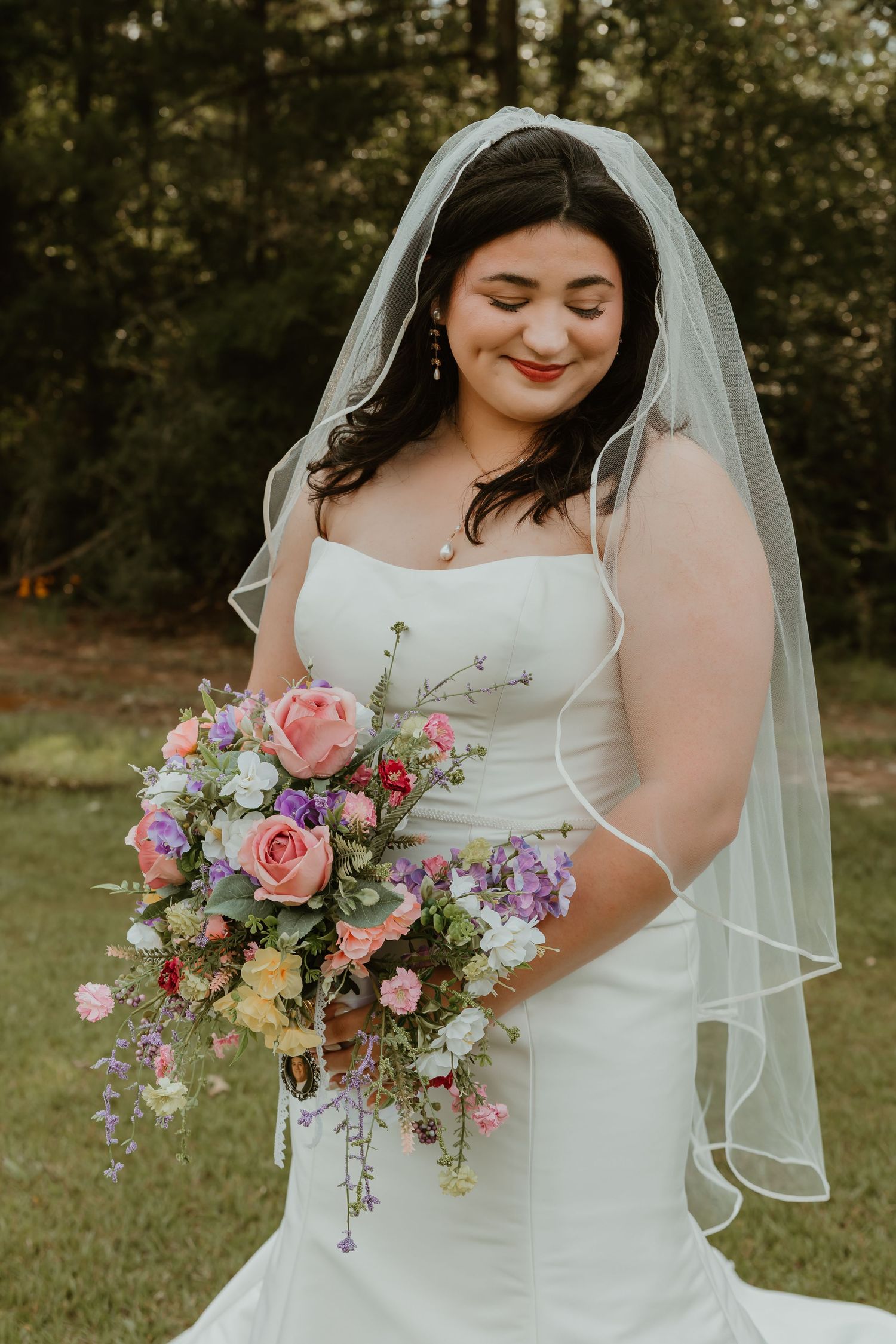 Bride in strapless gown and veil holds colorful cascading bouquet of roses and wildflowers outdoors.