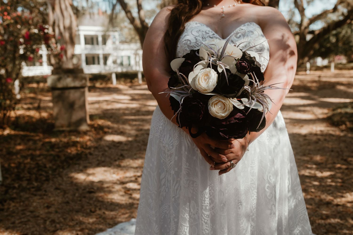 Bride in strapless lace gown holding a dark and white floral bouquet in front of a historic plantation home.