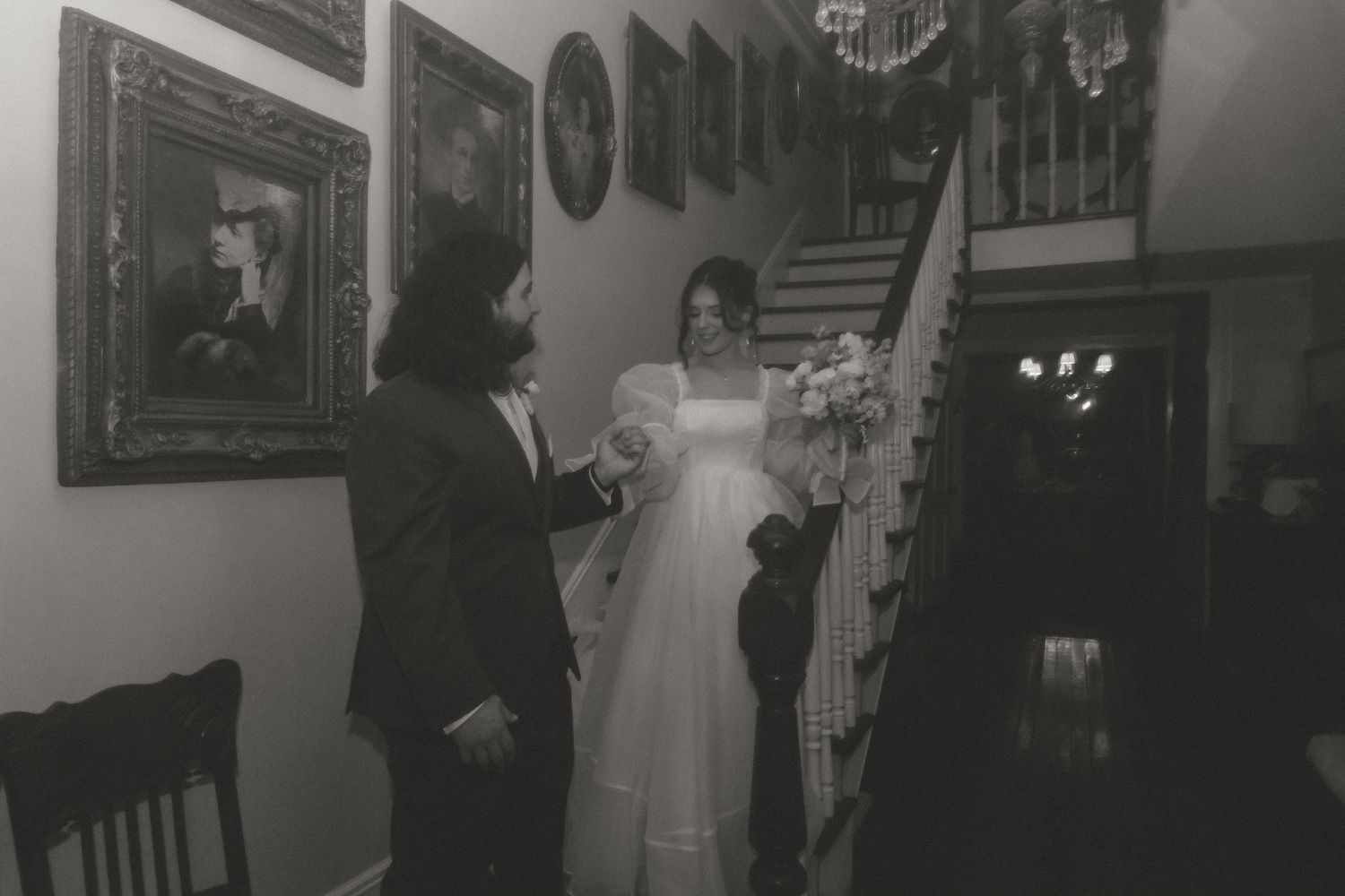 Black and white photo of bride descending staircase holding bouquet while groom watches inside historic home.