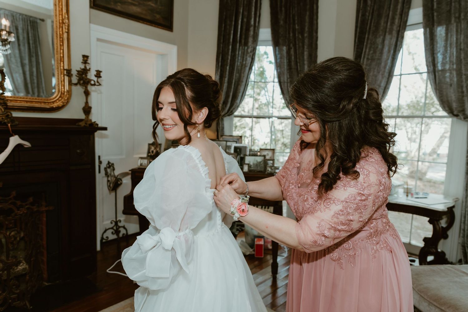Mom carefully fastening the back of a brides white wedding gown indoors.