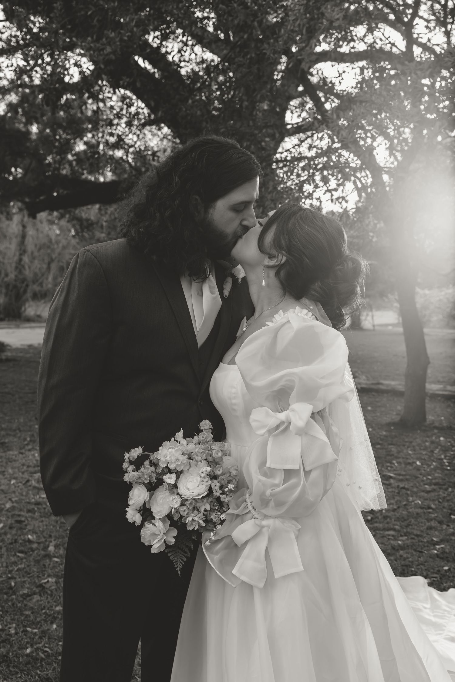 Black and white photo of bride and groom sharing first kiss under sunlit oak tree.