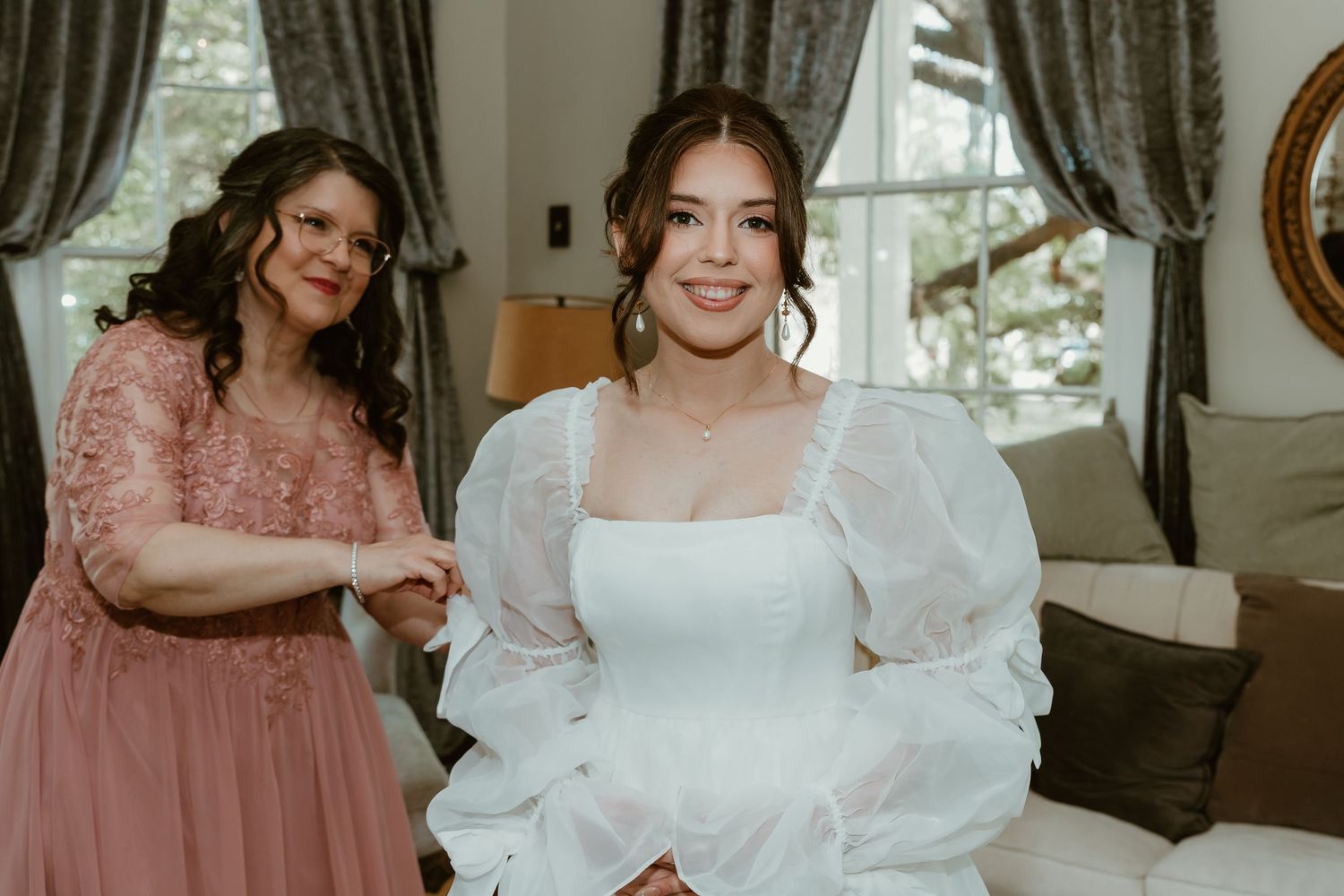 Smiling bride in white puff sleeve wedding gown holding hands with mother in pink lace dress.
