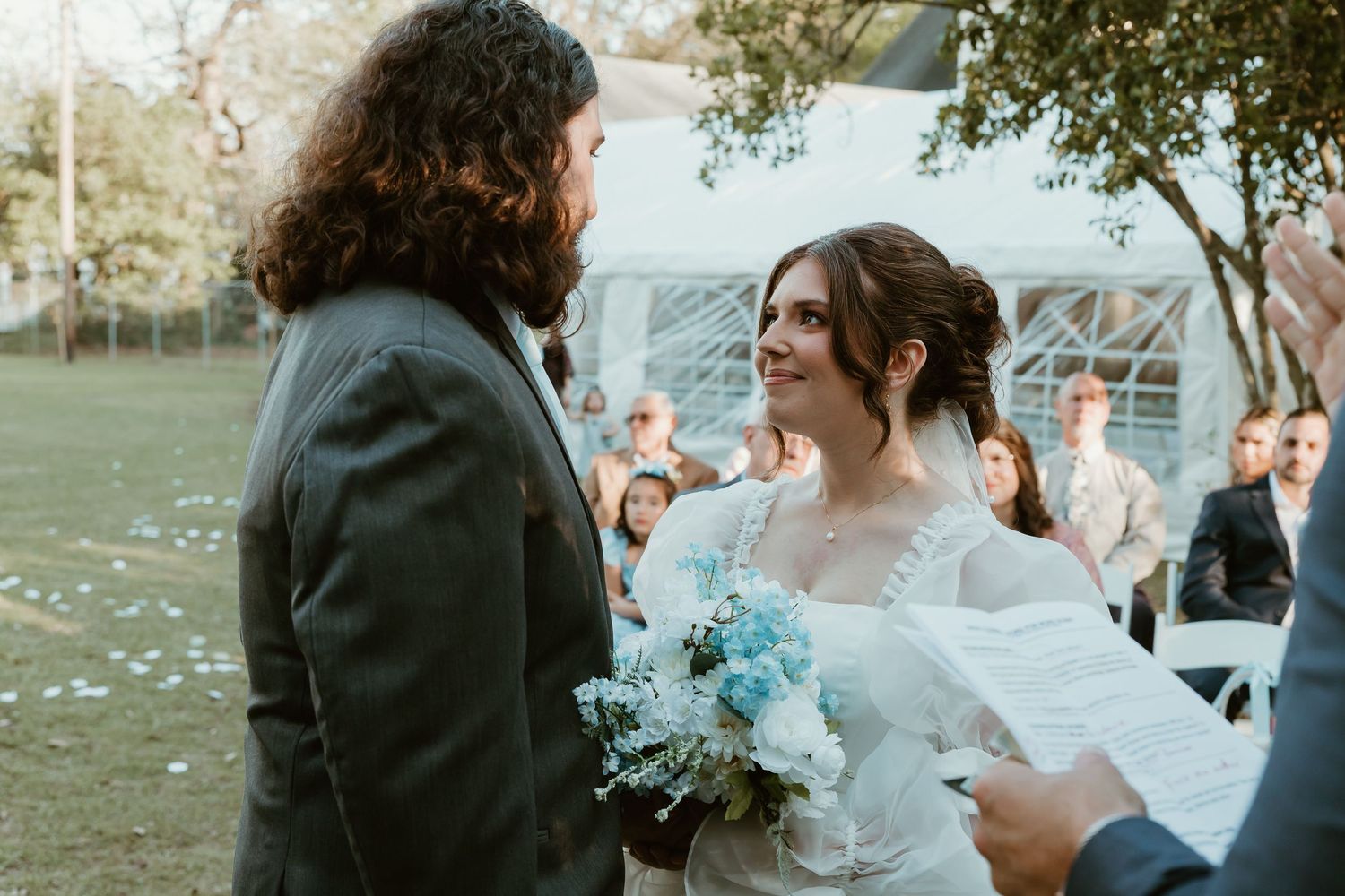 A smiling bride holding blue and white flowers gazes at her long-haired groom during an outdoor garden wedding ceremony.