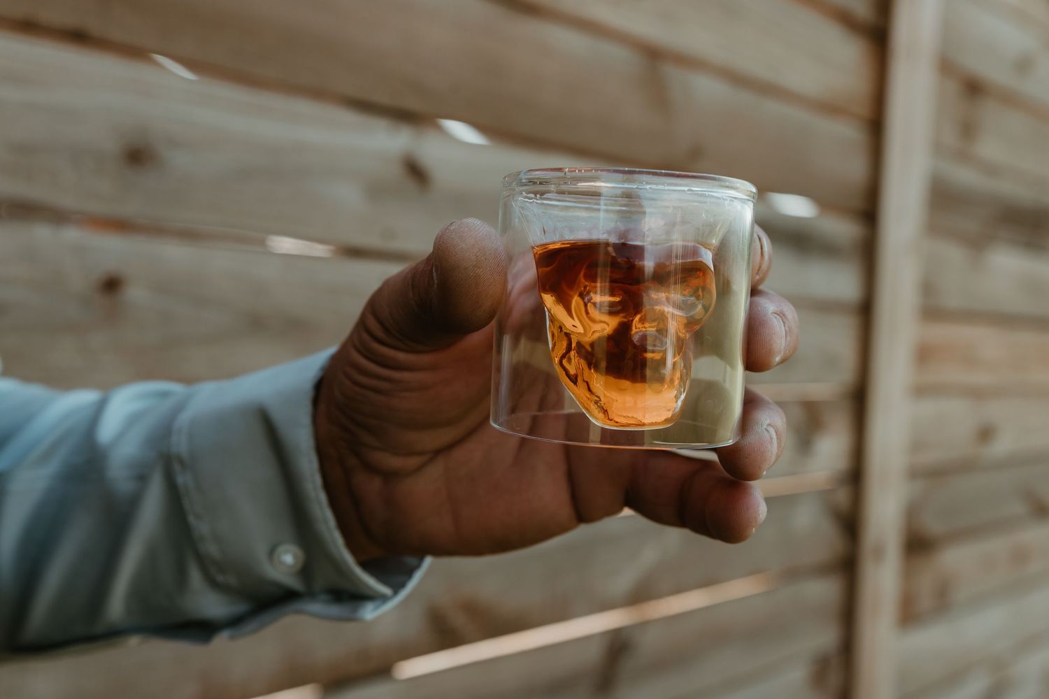Hand holding a skull-shaped whiskey glass with whiskey against a wooden background.
