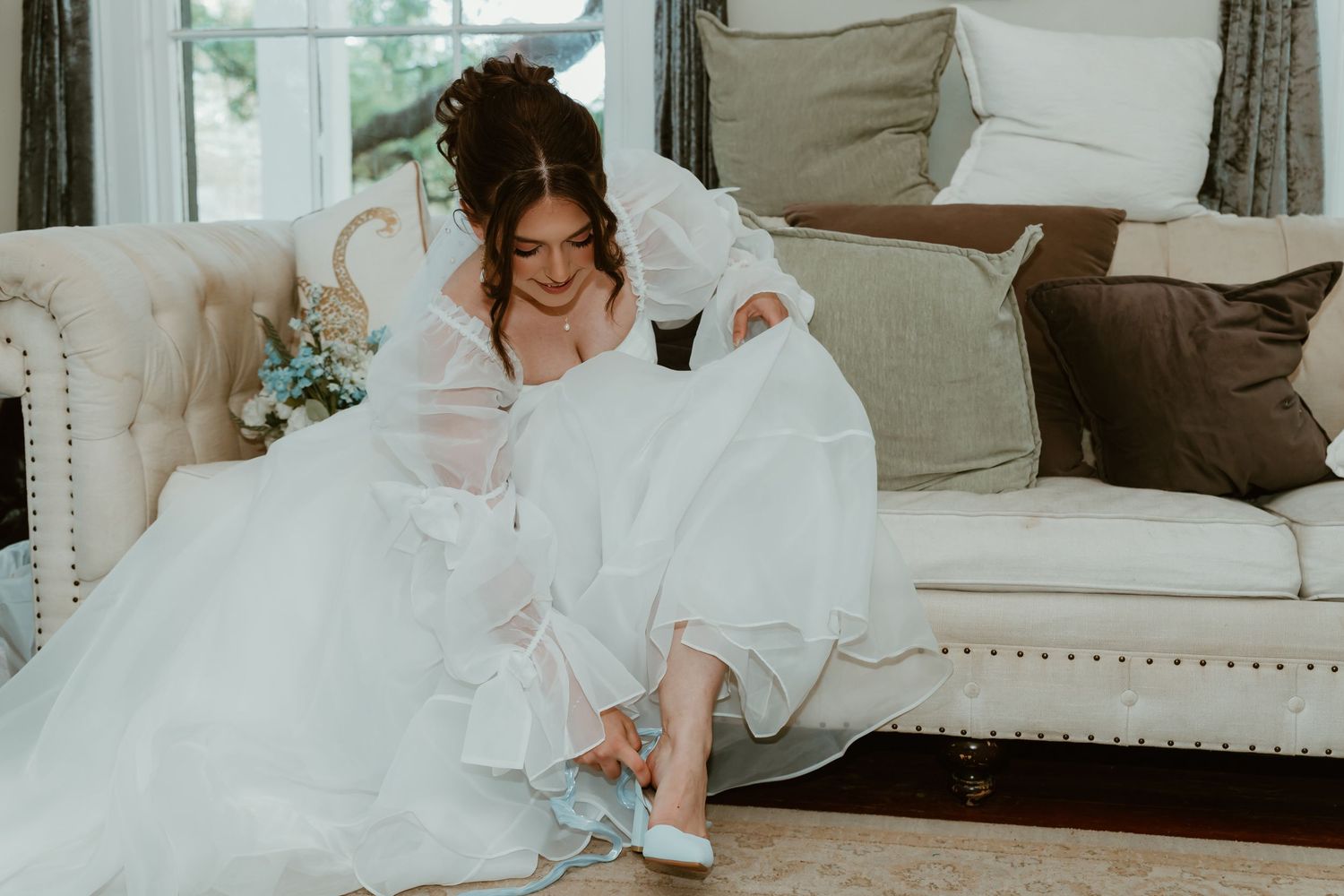 Bride in elegant white wedding dress admiring her something blue shoes while seated on a cream sofa.
