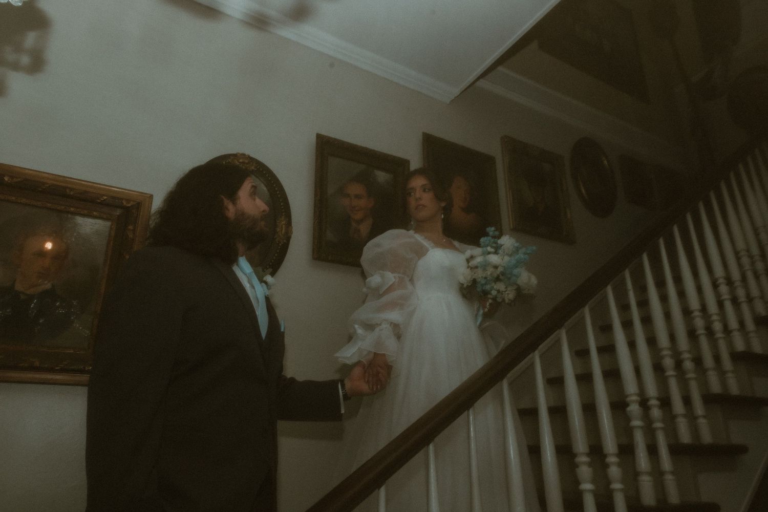 Bride descends staircase in white gown holding bouquet as groom waits in historic home.
