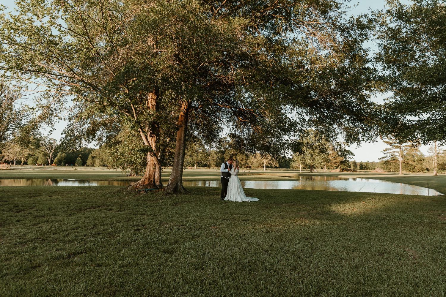 Bride and groom stand beneath a large oak tree beside a serene pond during golden hour outdoor wedding.