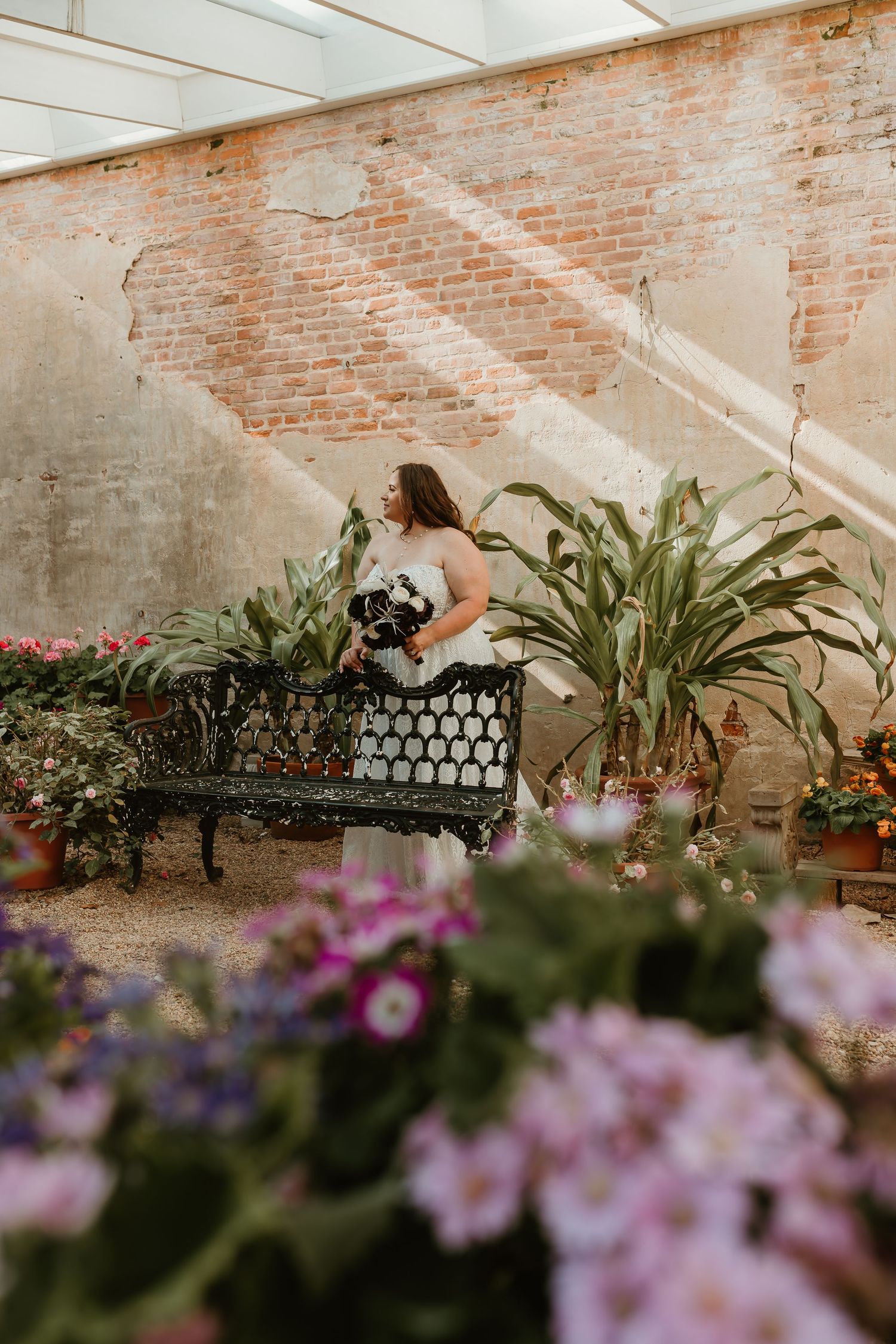 Bride in lace wedding gown sitting on vintage iron bench in greenhouse with brick wall and flowers.
