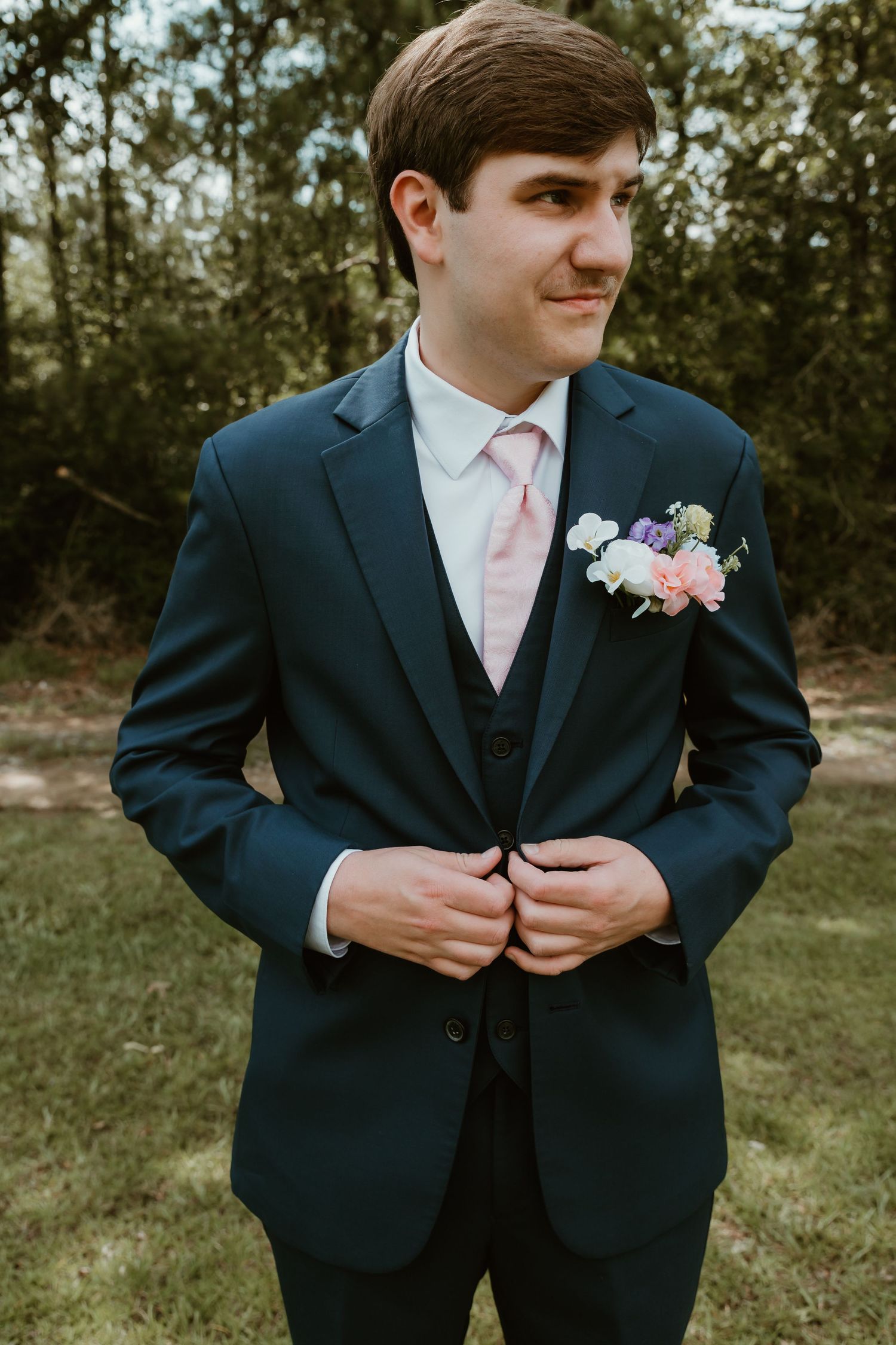 Groom in navy suit adjusts jacket button while wearing colorful floral boutonniere outdoors.