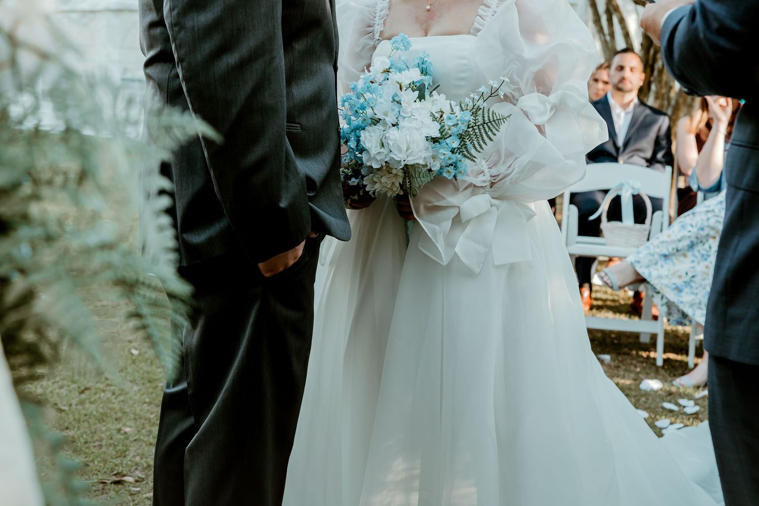 Bride holding blue and white bouquet stands with groom during outdoor wedding ceremony.