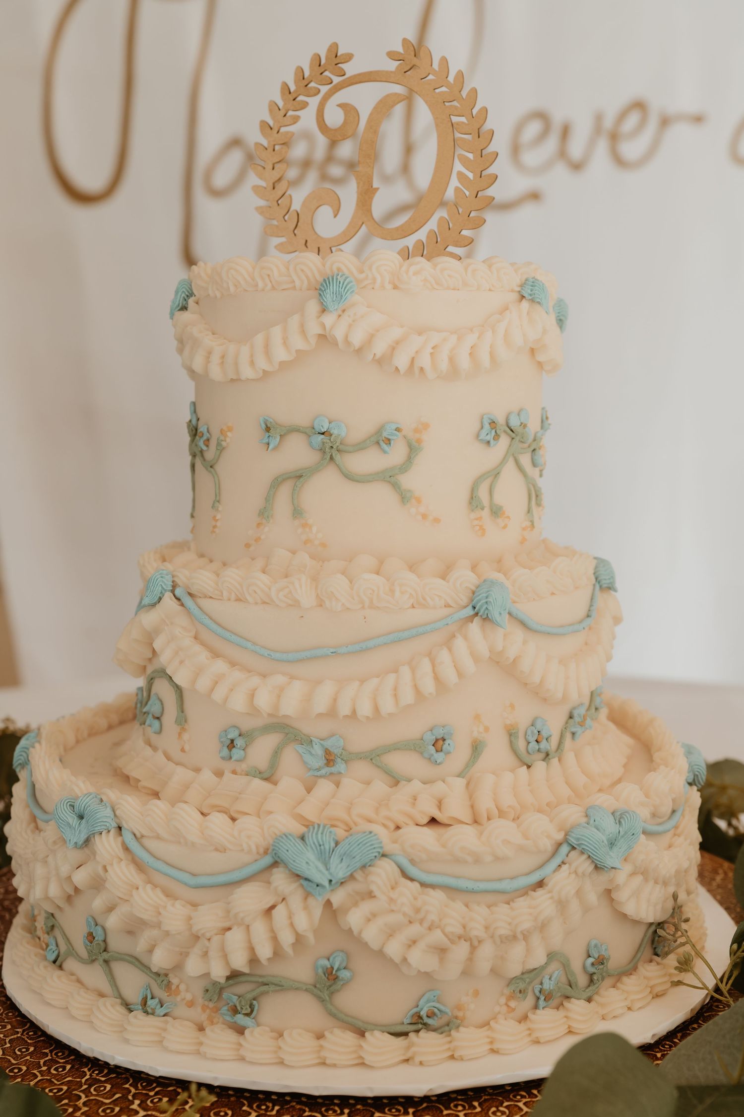 Three-tier white wedding cake with blue bow details and gold monogram topper on display.