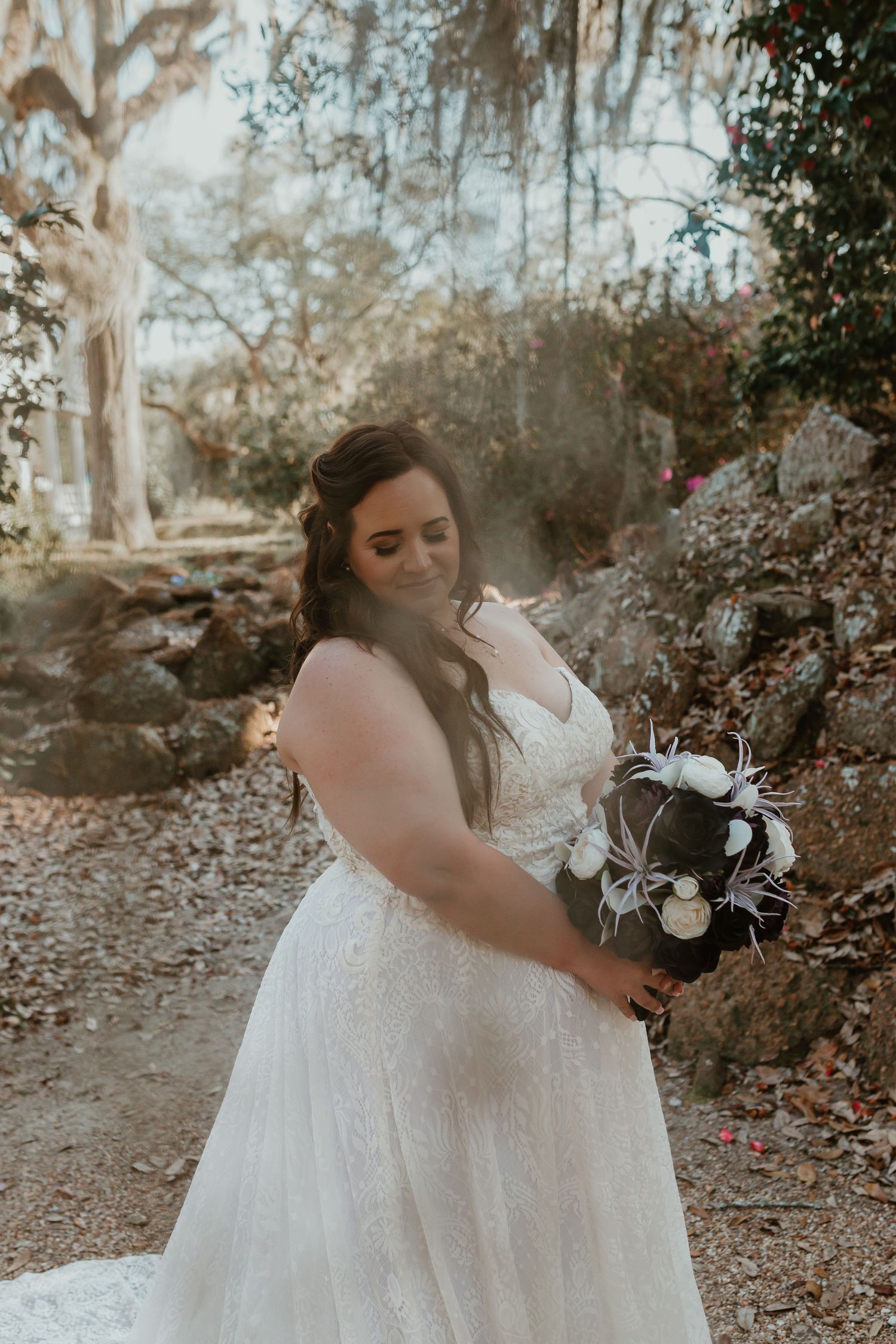 Bride in lace wedding gown holding dark floral bouquet with feathers in wooded garden setting.