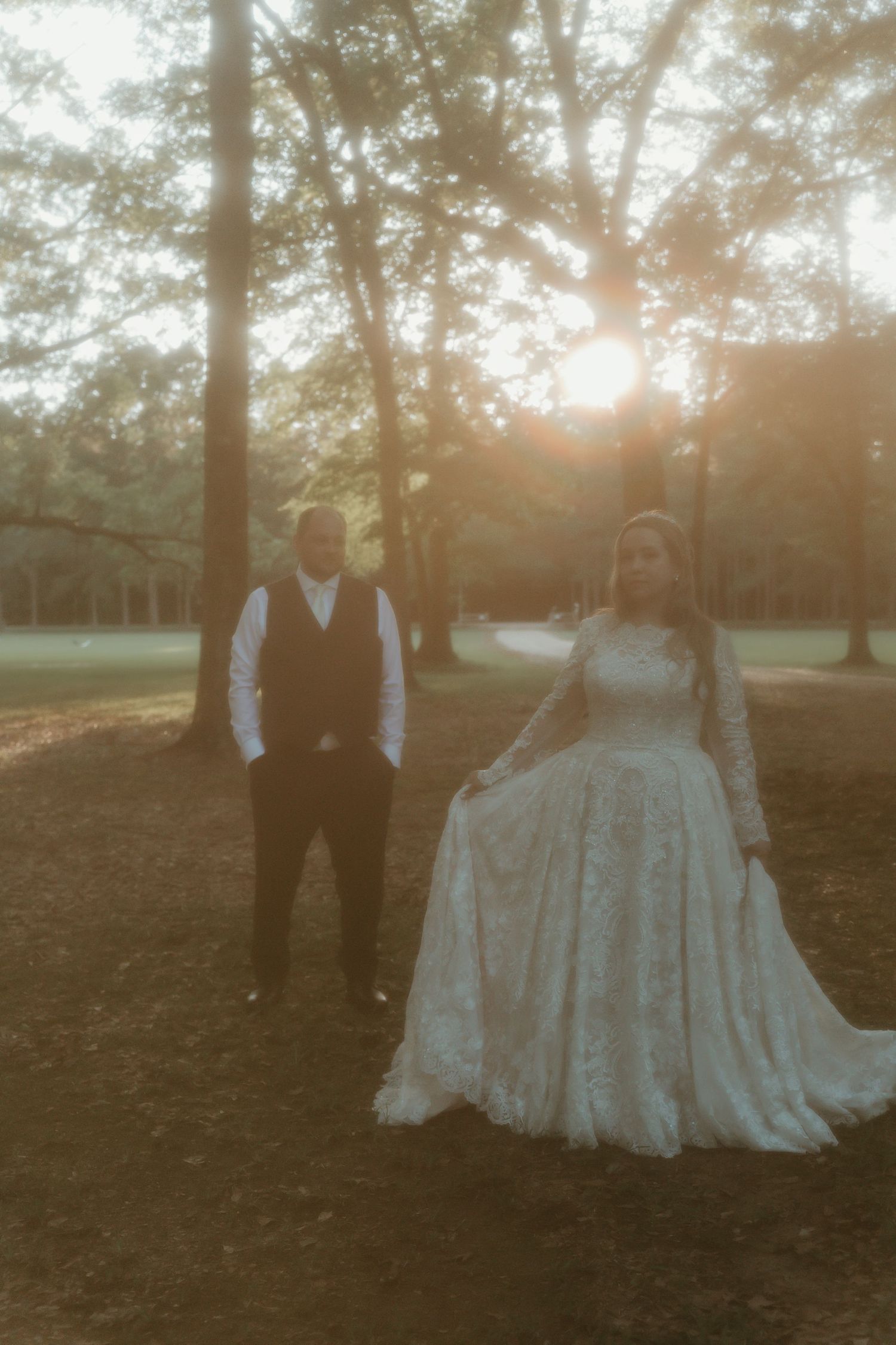Bride in long-sleeve lace gown and groom stand among tall trees bathed in warm golden sunset light.