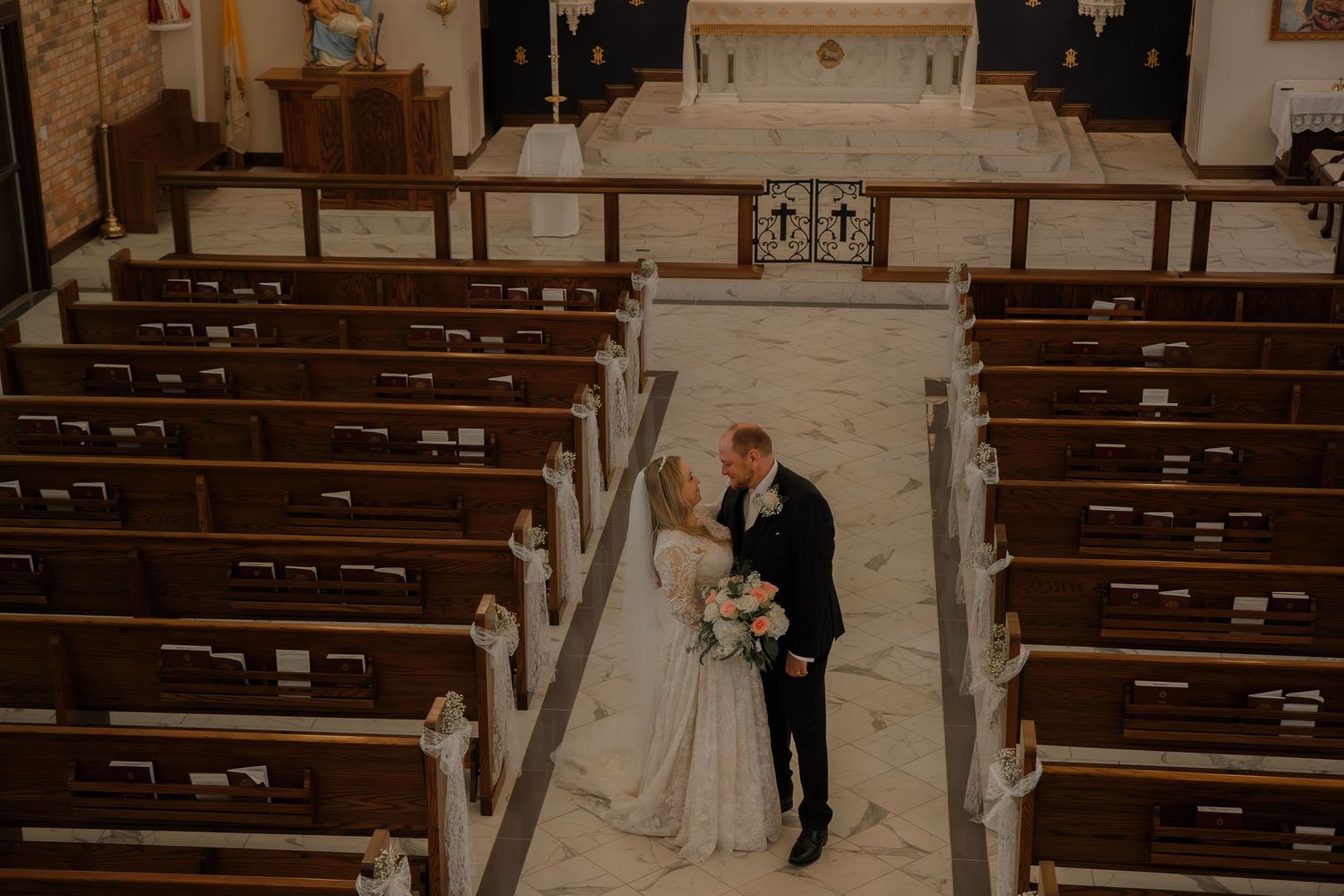 Bride and groom share a romantic moment in the aisle of a traditional Catholic church with wooden pews.