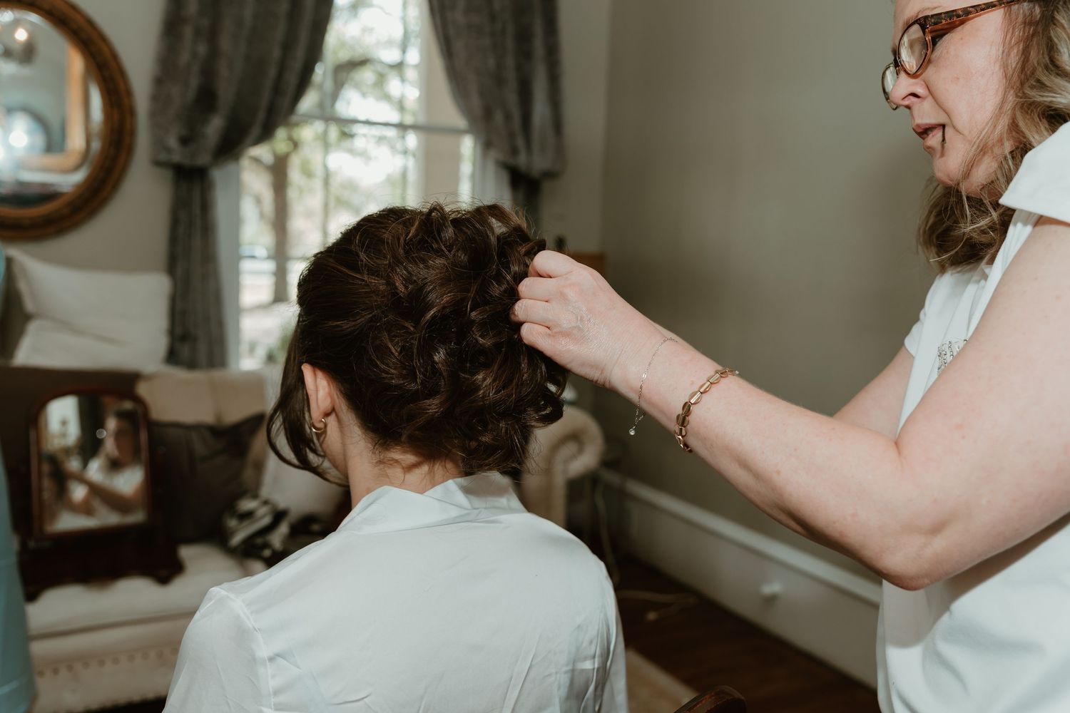 Hairstylist pinning an elegant updo on a bride seated near a window in a bright dressing room.