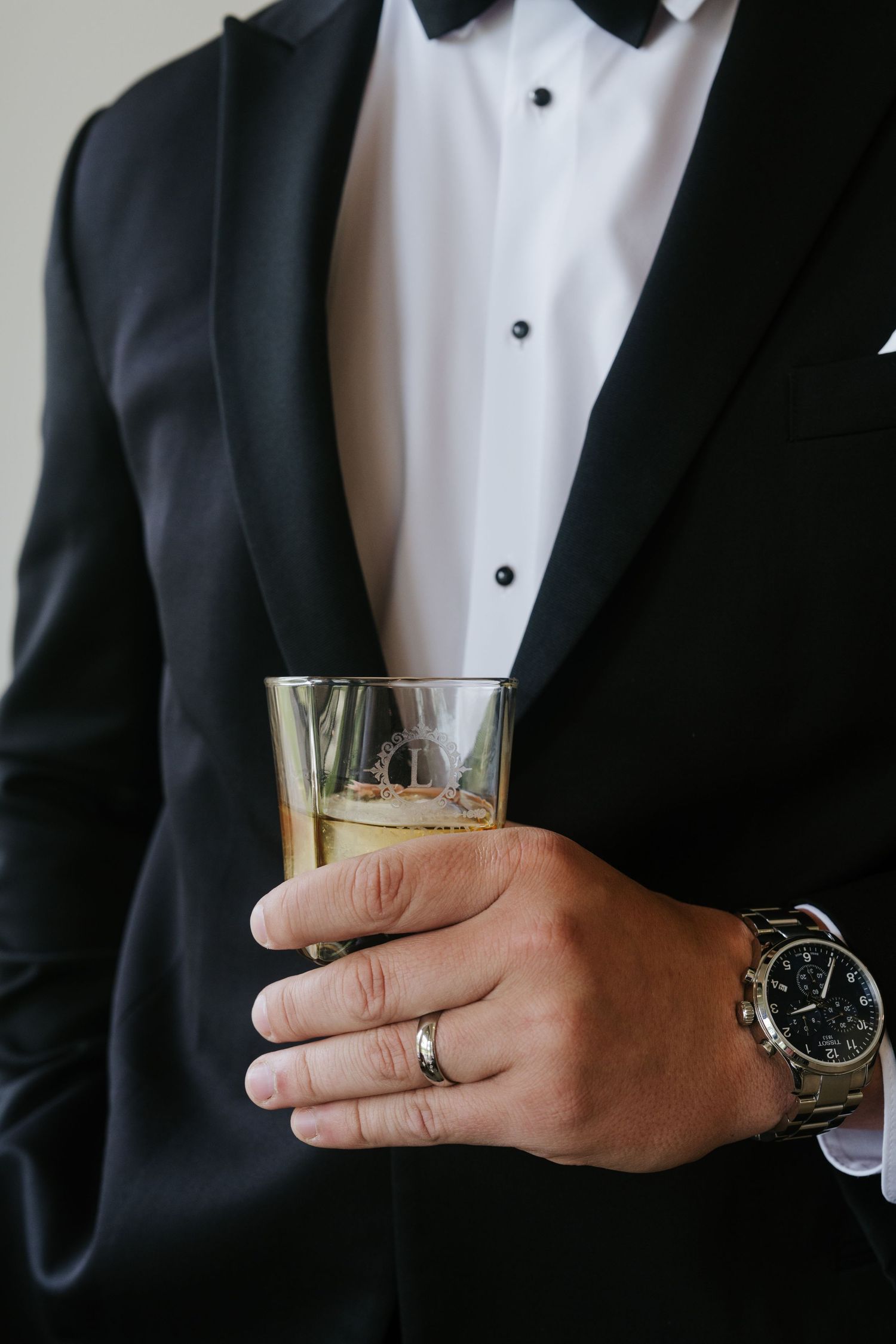Close-up of a hand holding a champagne glass wearing a black tuxedo jacket, silver watch and wedding ring.