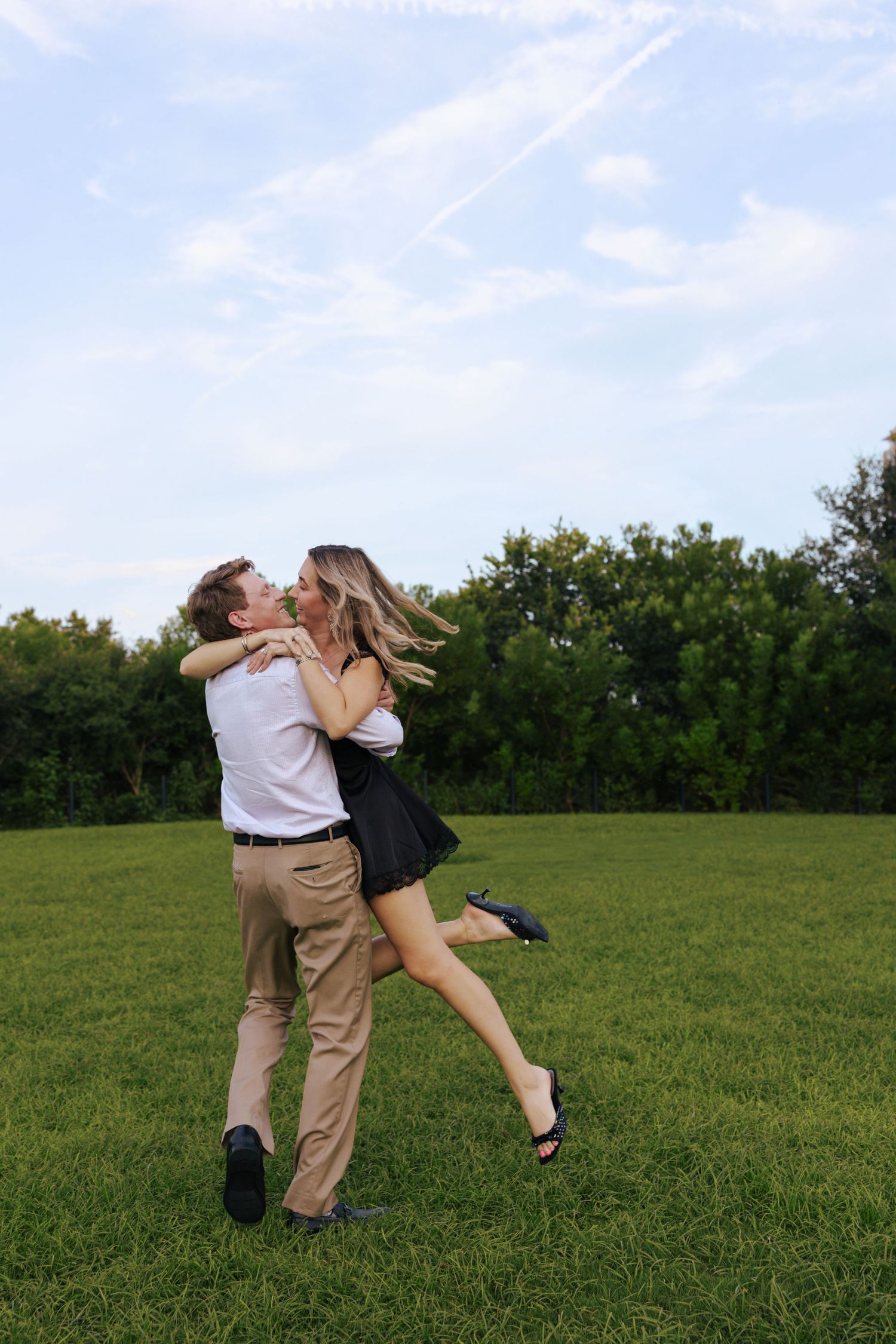 A romantic couple embraces and kisses while standing in a green grassy field on a summer day.