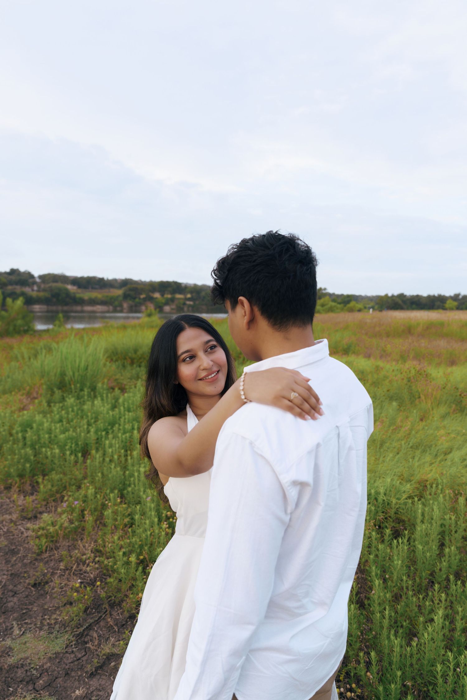 A couple embraces in a scenic field with tall grass and natural landscape during a romantic outdoor photoshoot.