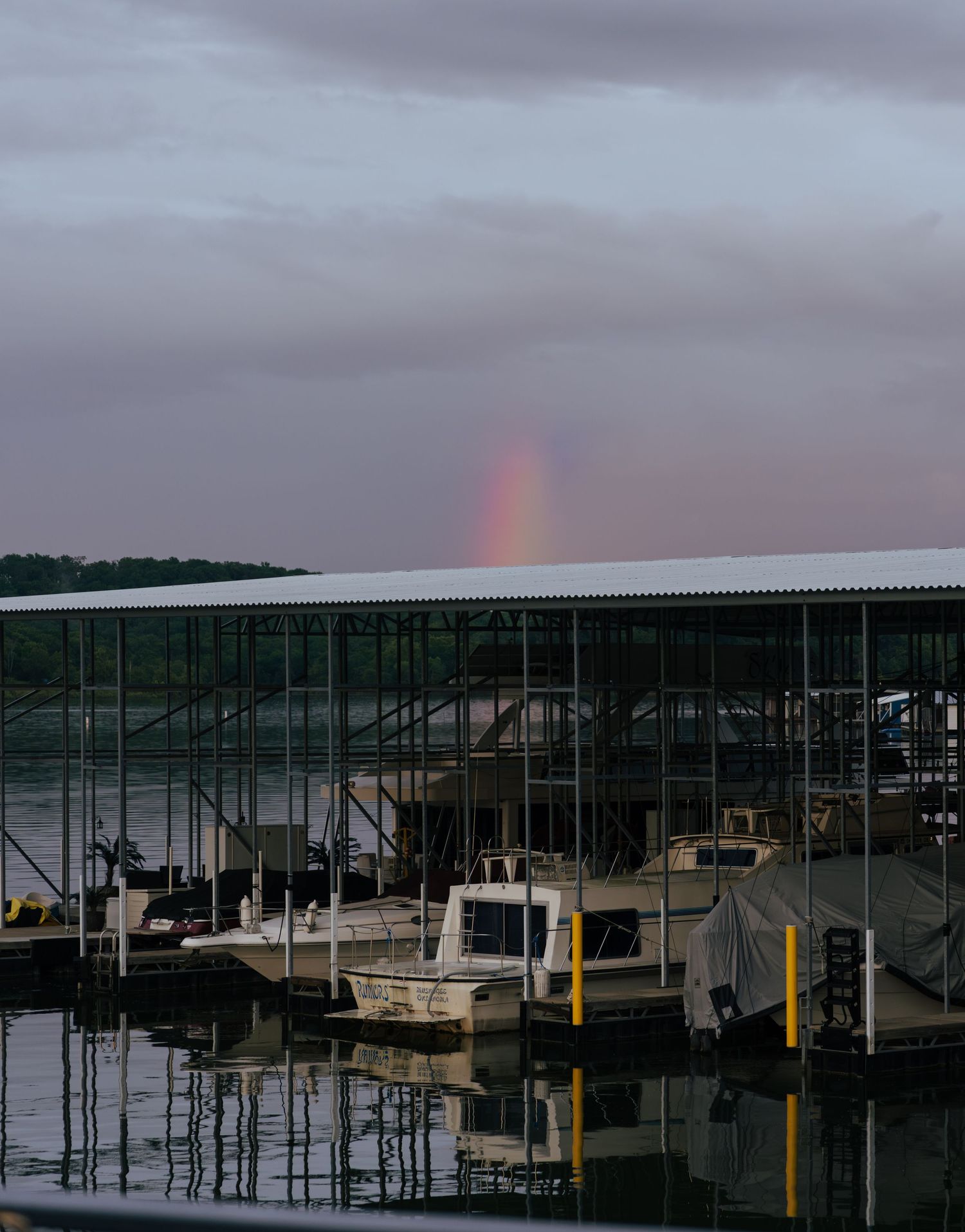 A rainbow appears over a marina with covered boat slips and docked vessels under cloudy skies at dusk.