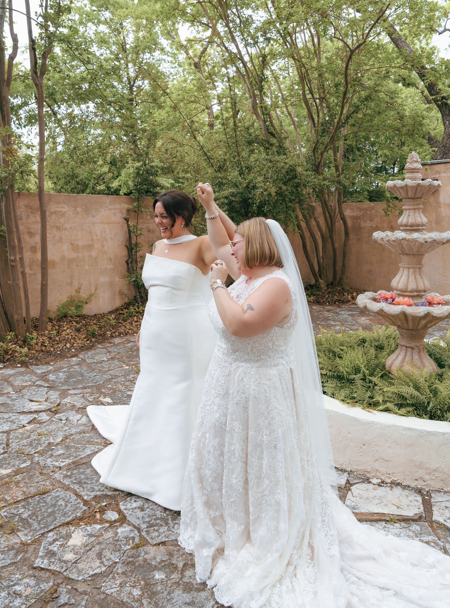 Two people in white wedding dresses dance together in a garden courtyard surrounded by trees and stone walls.