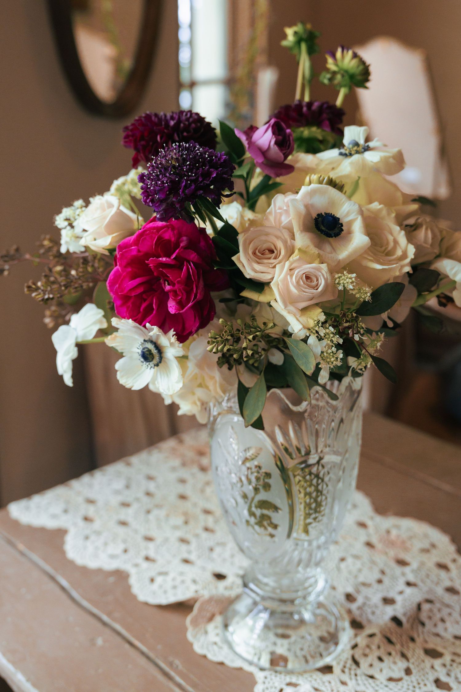 A vintage crystal vase holds an elegant arrangement of roses, anemones, and purple dahlias on a lace doily.