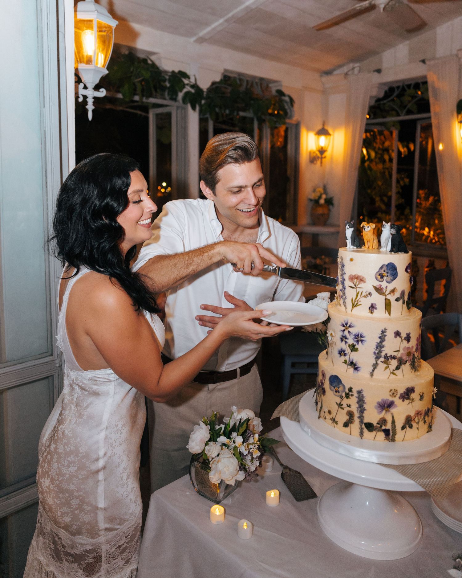 Bride and groom smiling while cutting a beautiful three-tier floral wedding cake at their elegant evening reception.