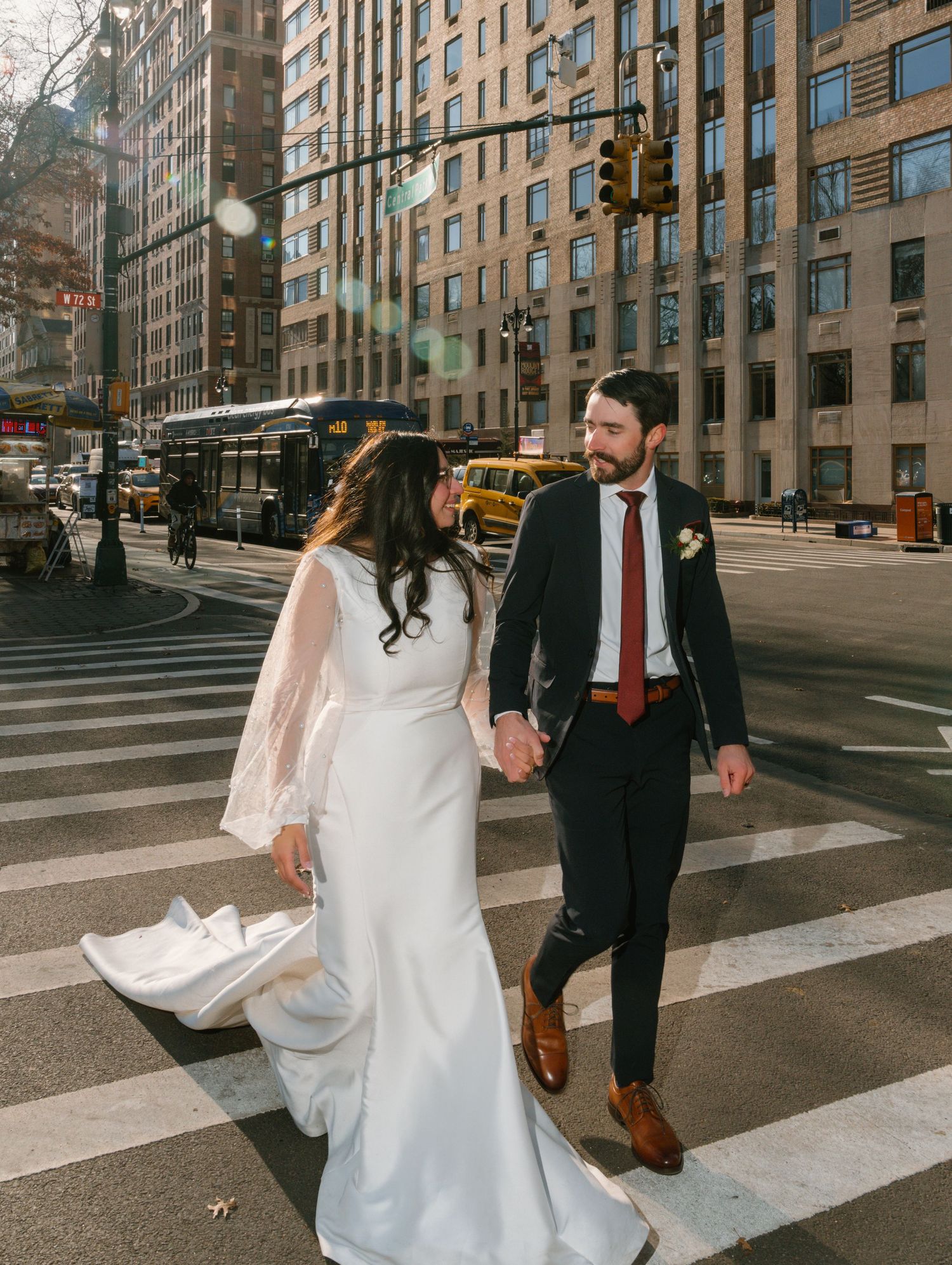 Newlyweds walking across urban crosswalk in New York City with tall buildings and yellow taxi in background.