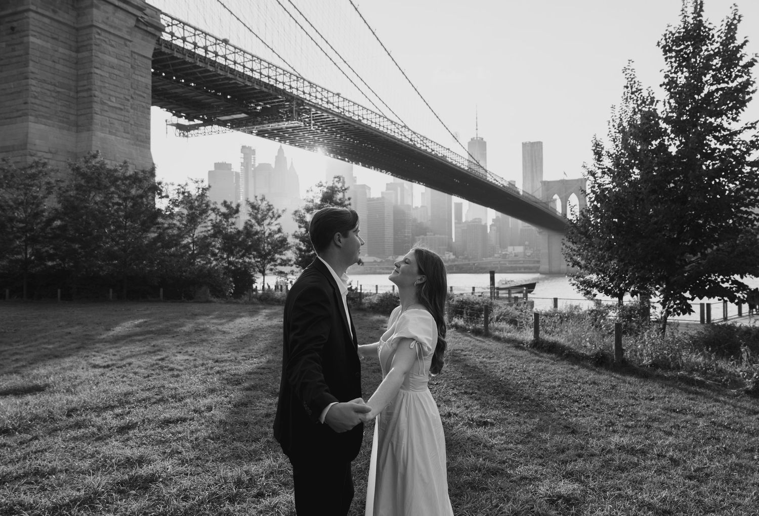 Romantic couple poses beneath Brooklyn Bridge in black and white photograph.