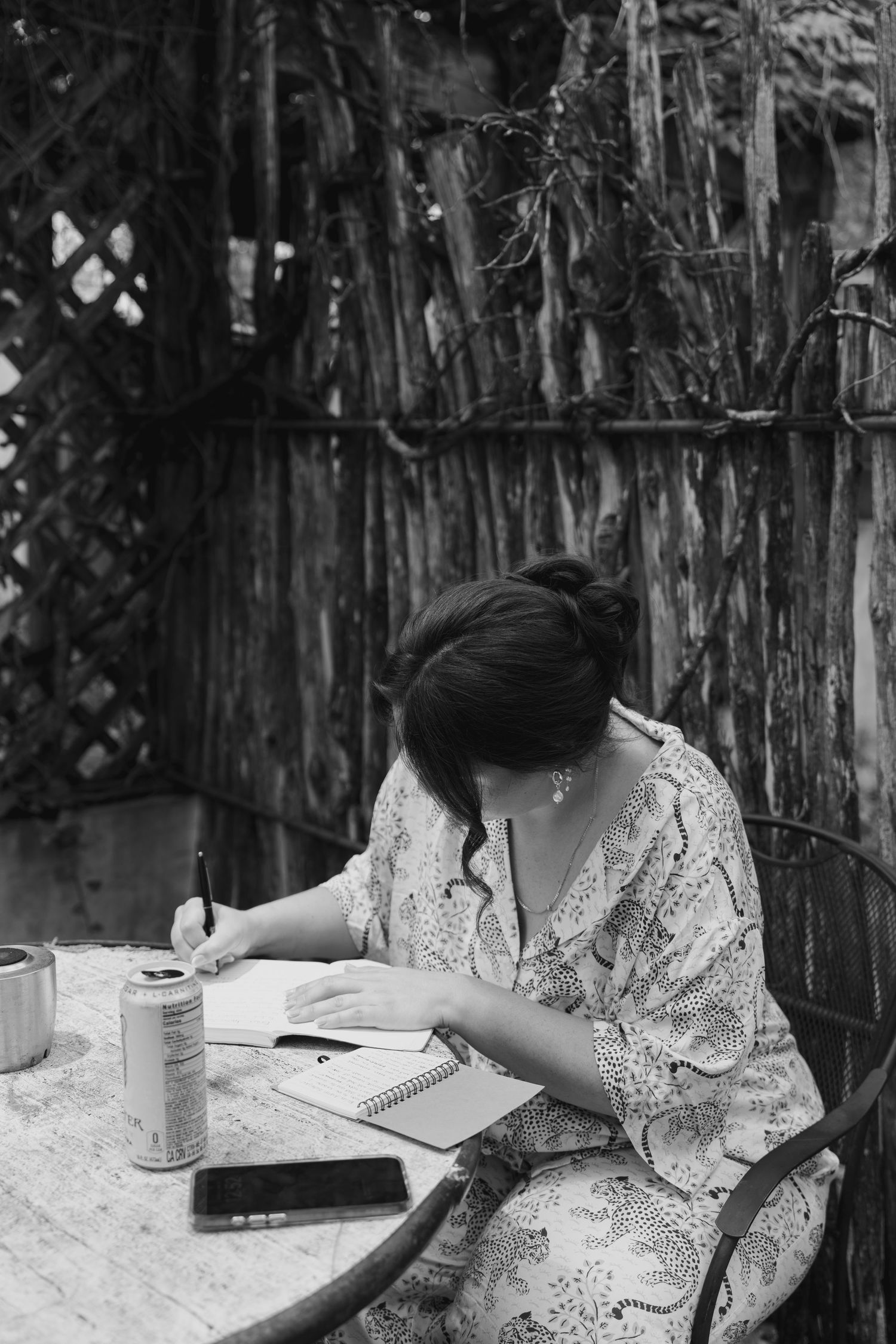 A person in a patterned shirt writes in a notebook at a table in black and white photography.