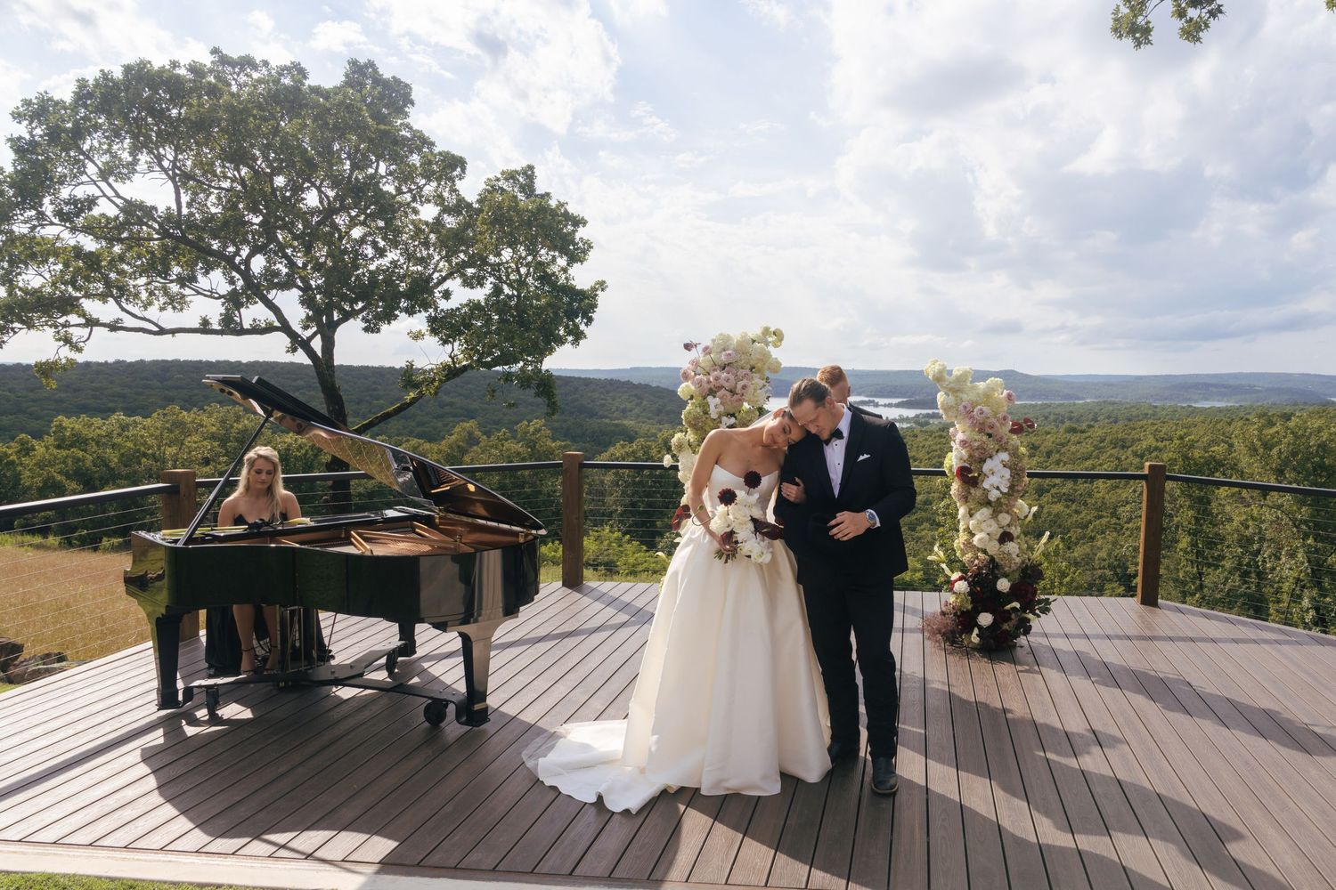 A wedding ceremony takes place on a scenic deck overlooking mountains and lakes with elegant floral arrangements.