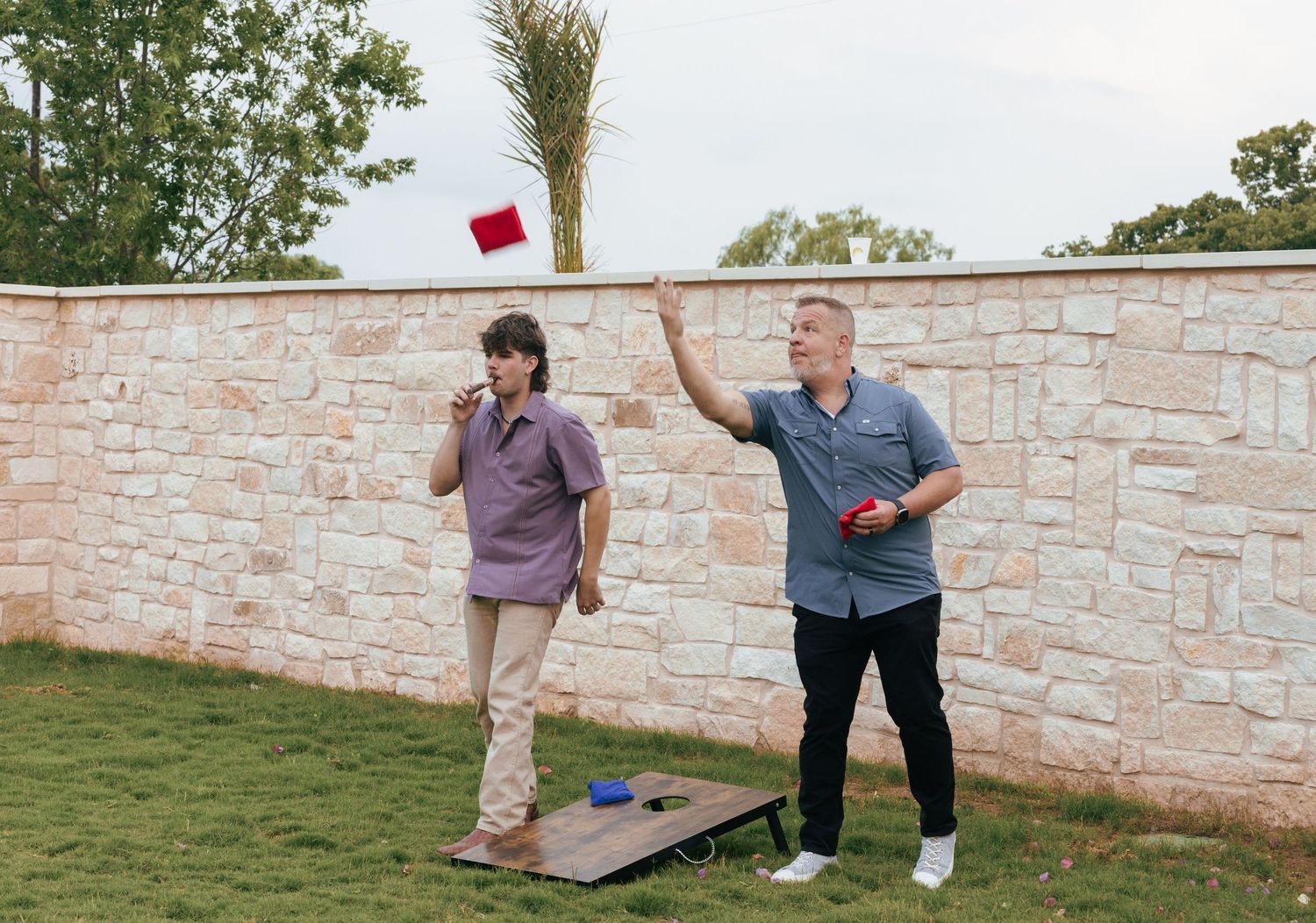 Two people play cornhole on a lawn next to a stone wall during a casual outdoor game.