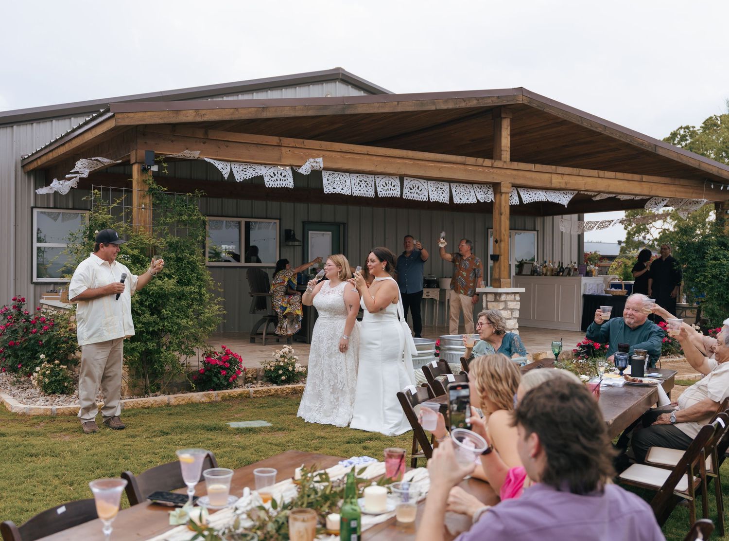 Wedding guests gather outdoors under a wooden pergola for a celebration event at sunset.