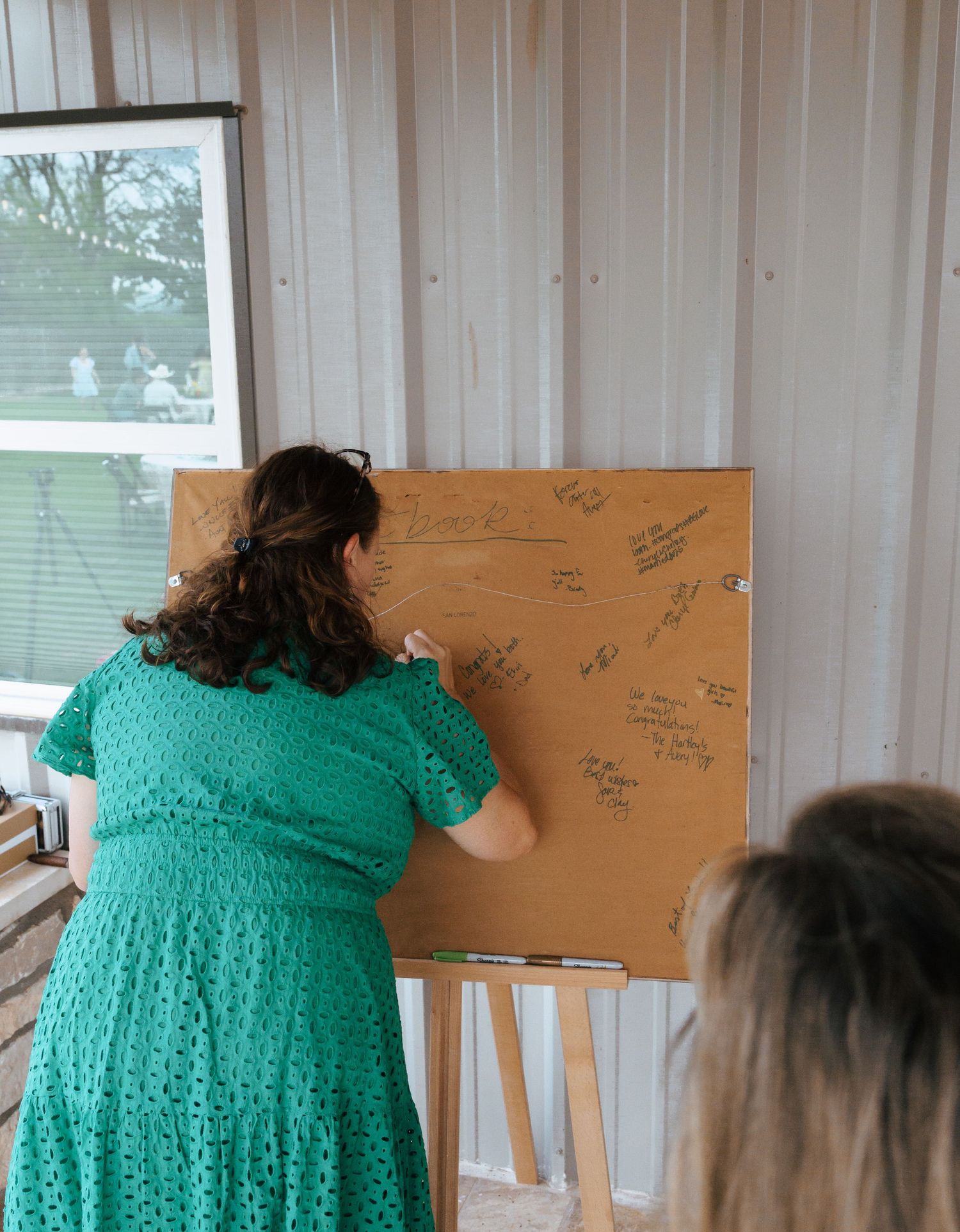 Someone in a turquoise dress writes on a large brown paper sheet attached to a wall in a room with windows.