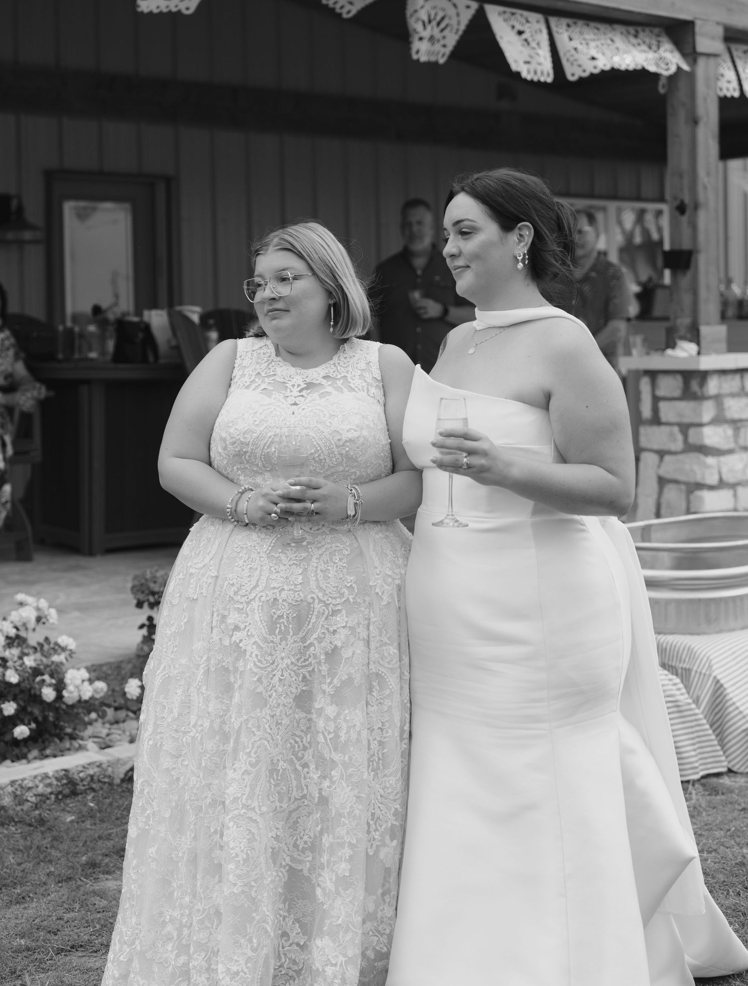 Two people in white formal dresses stand together during a wedding celebration outdoors.