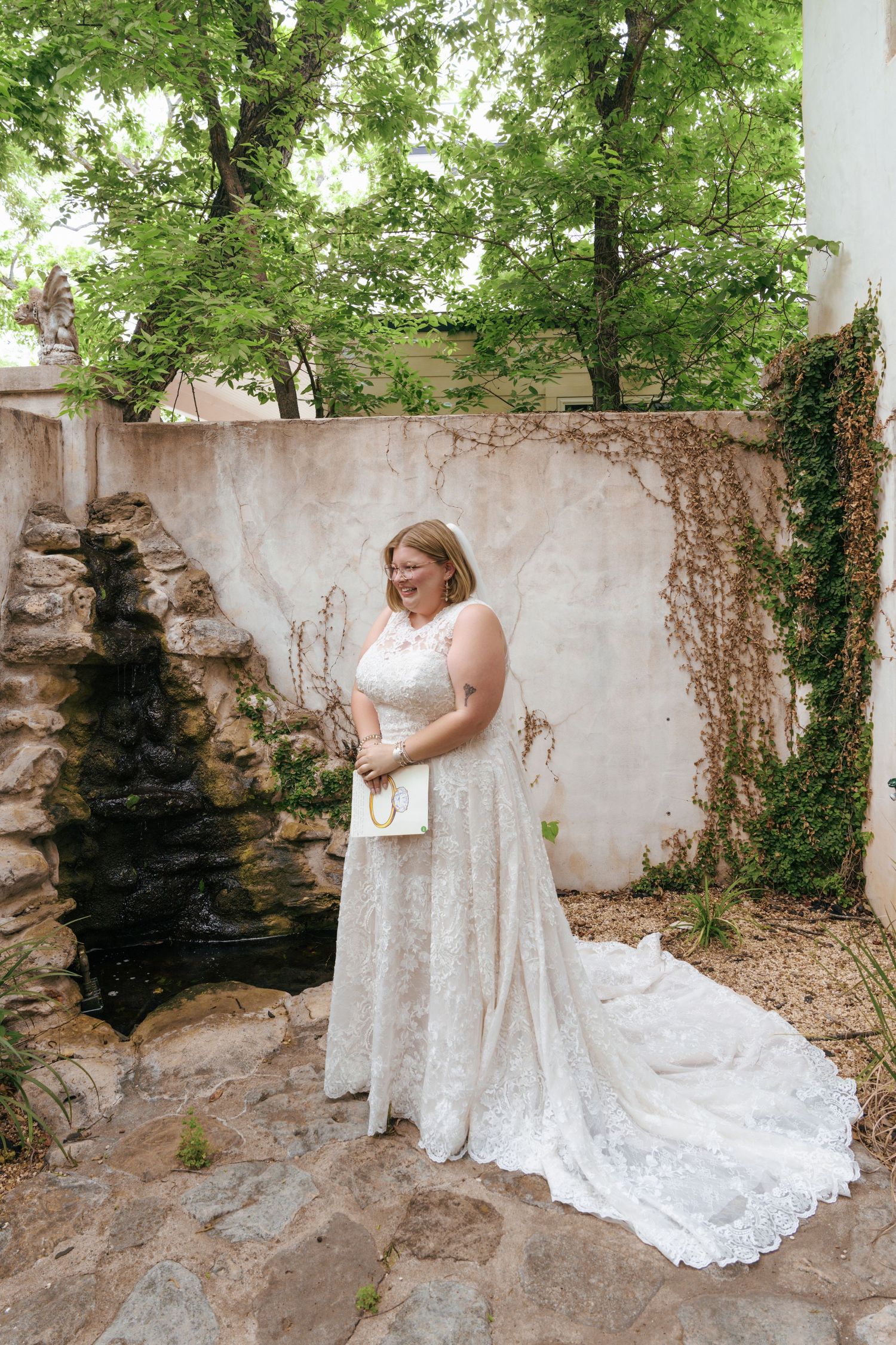 White lace wedding dress with long train photographed in a rustic stone wall setting with green foliage.
