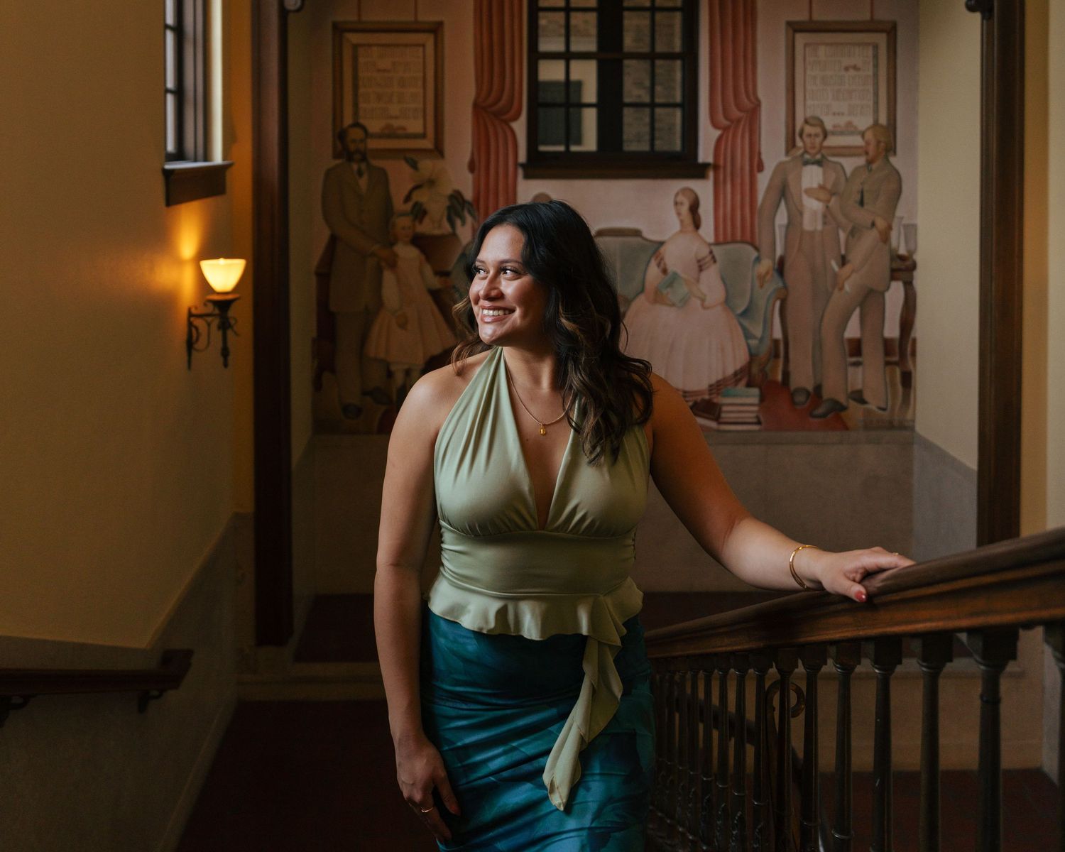 Woman in teal dress poses in elegant hallway with warm lighting and architectural details.