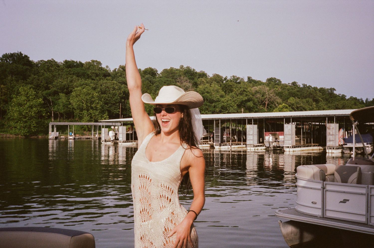 Person in floral dress poses near waterfront dock with boats and trees visible in the background.